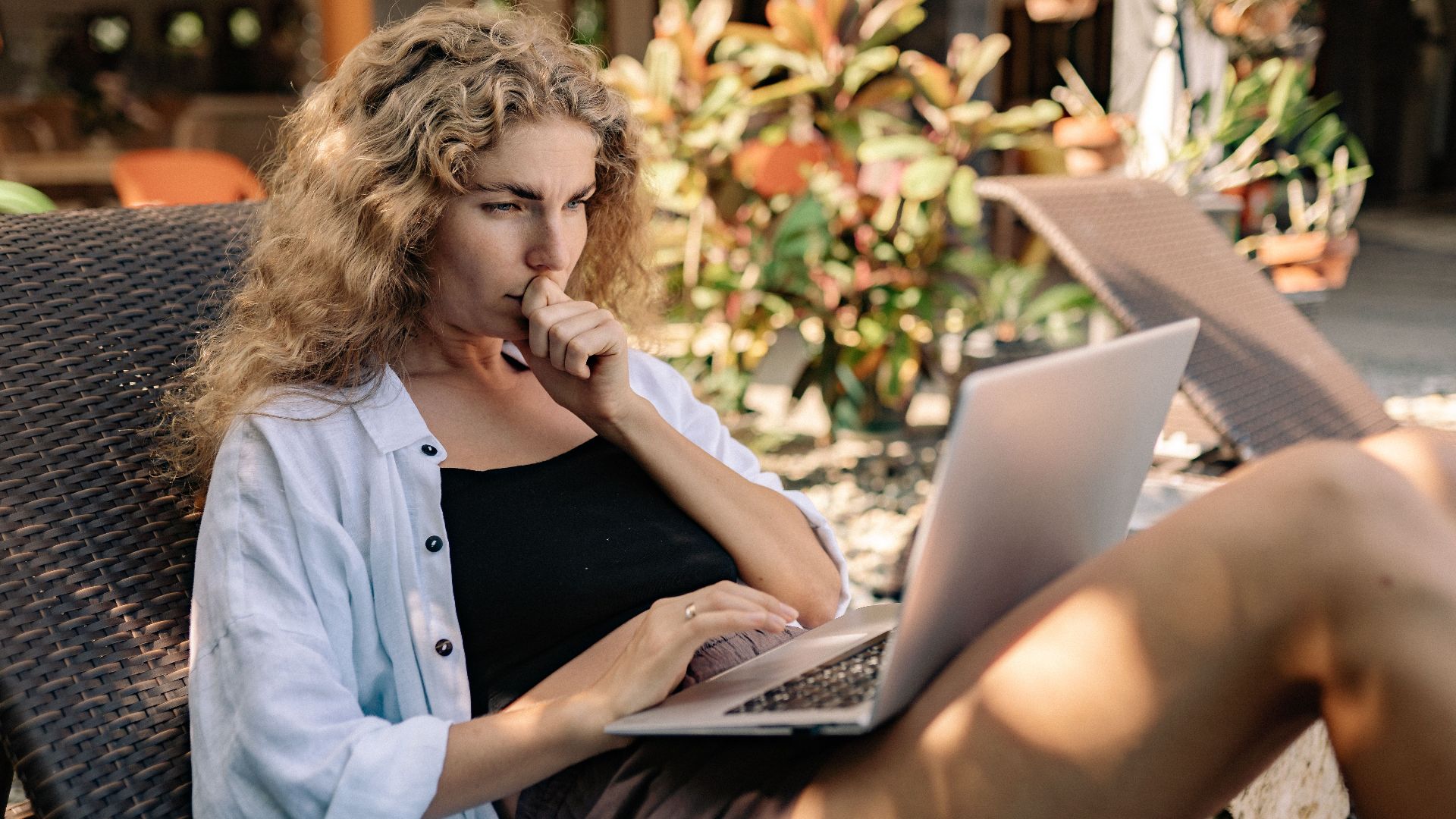 Woman with curly hair working remotely on a laptop while lounging outdoors in a tropical setting.
