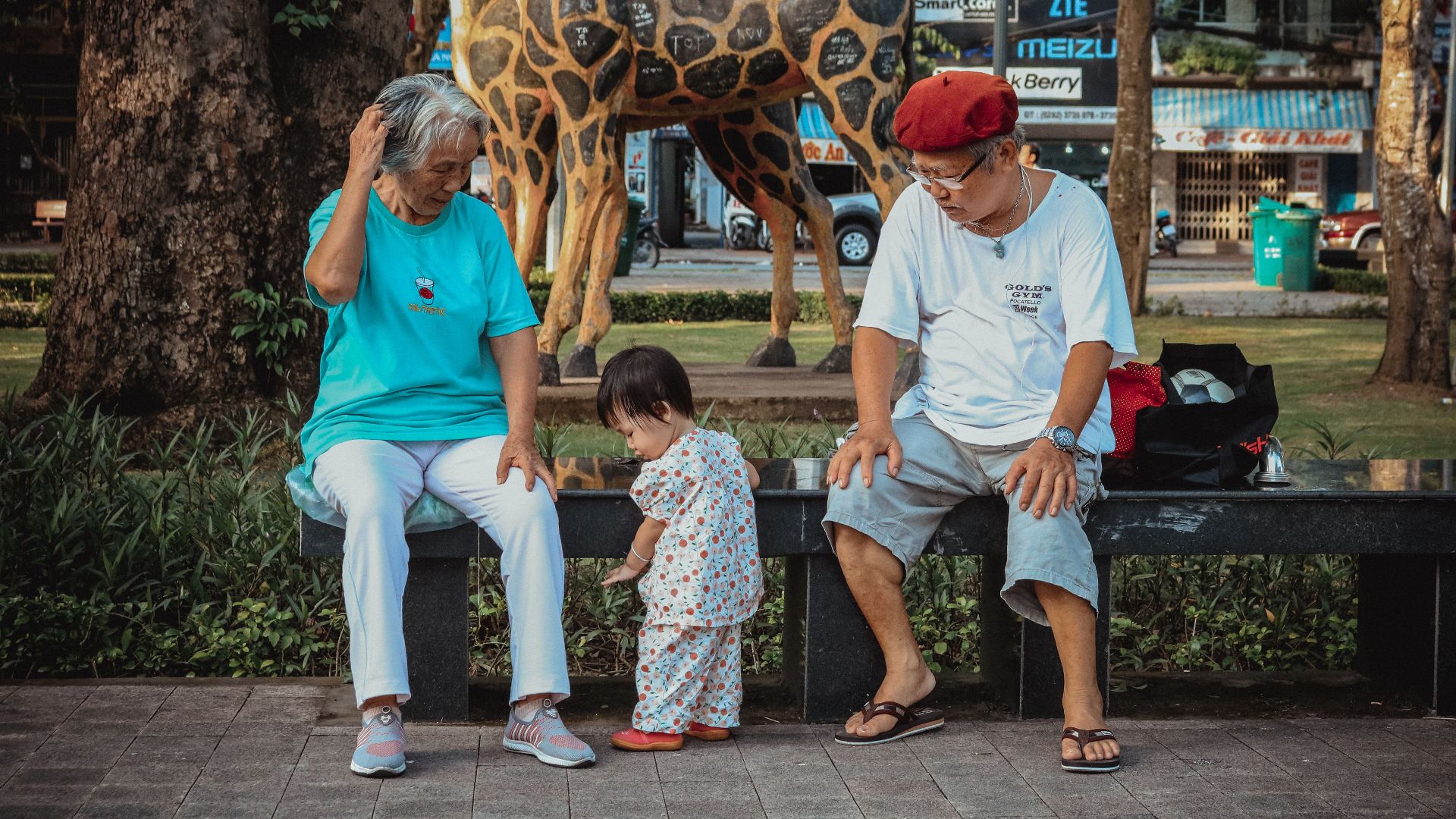 Elderly couple with toddler sitting on a bench in a Vietnamese park with unique giraffe sculpture.