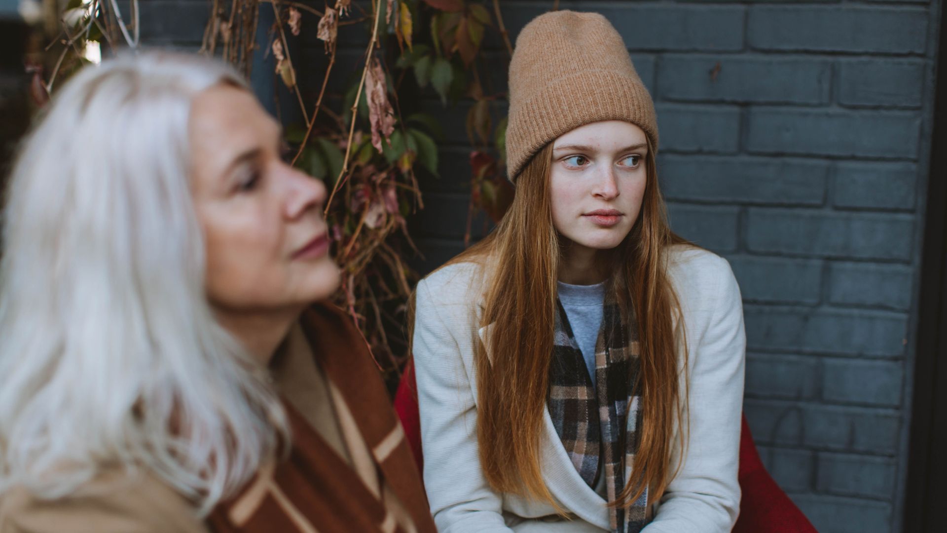 Two women sitting outdoors, wearing stylish autumn fashion, wool hats and scarves, against a brick wall.