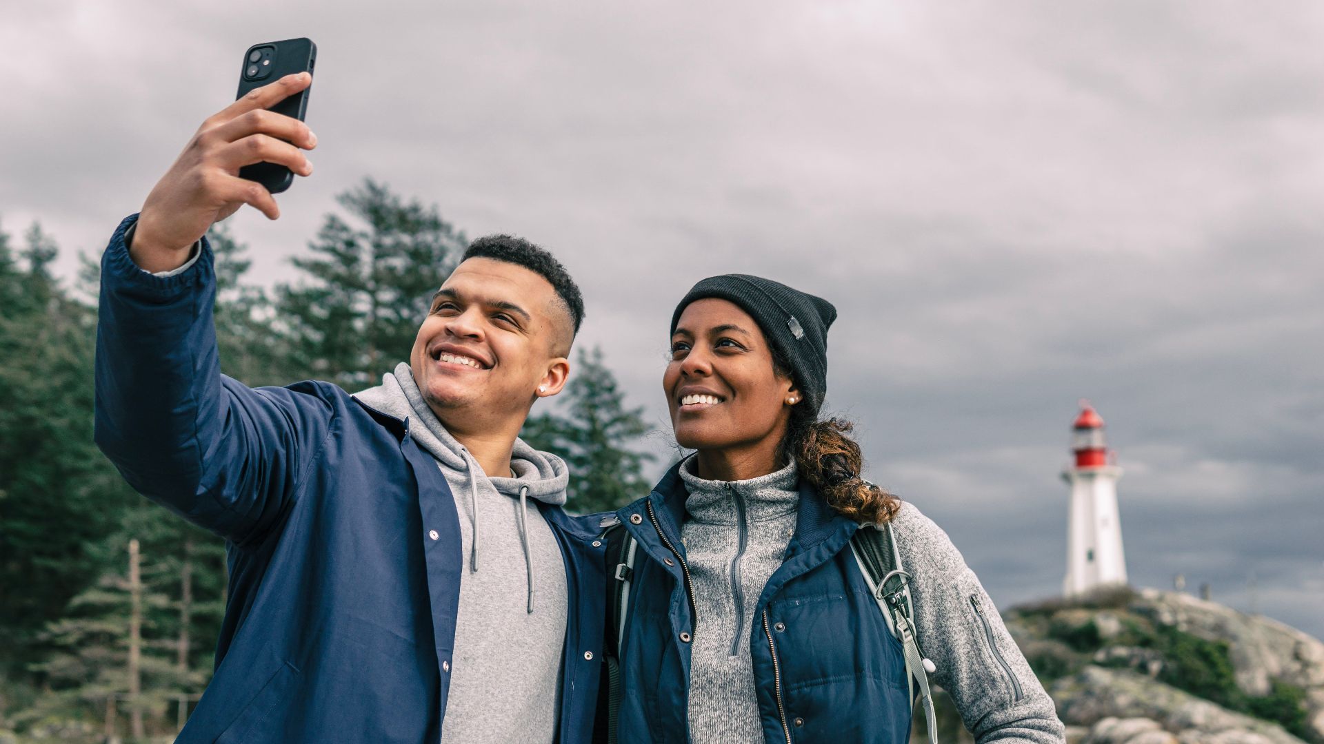 Happy couple taking a selfie with a scenic lighthouse in the background during a hiking adventure.