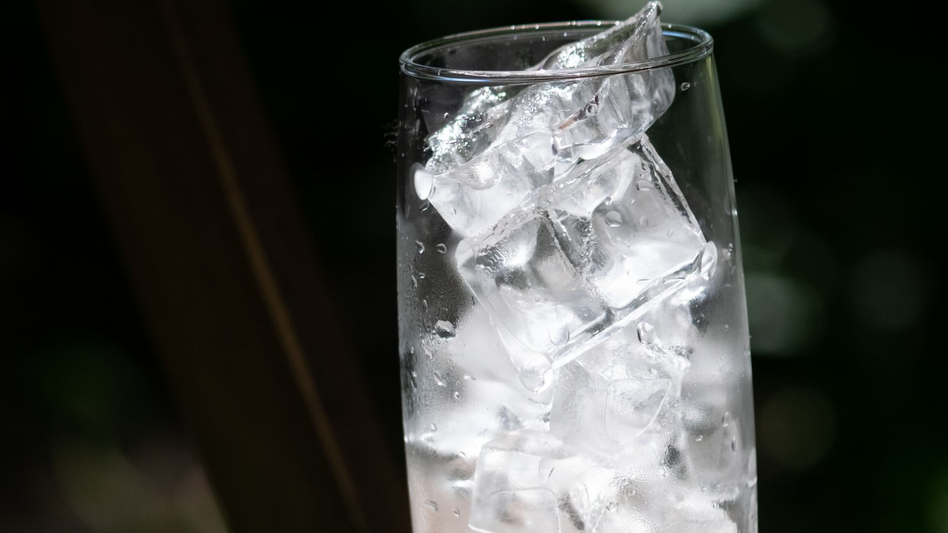 A tall glass filled with ice cubes resting on a wooden table outdoors in sunlight.