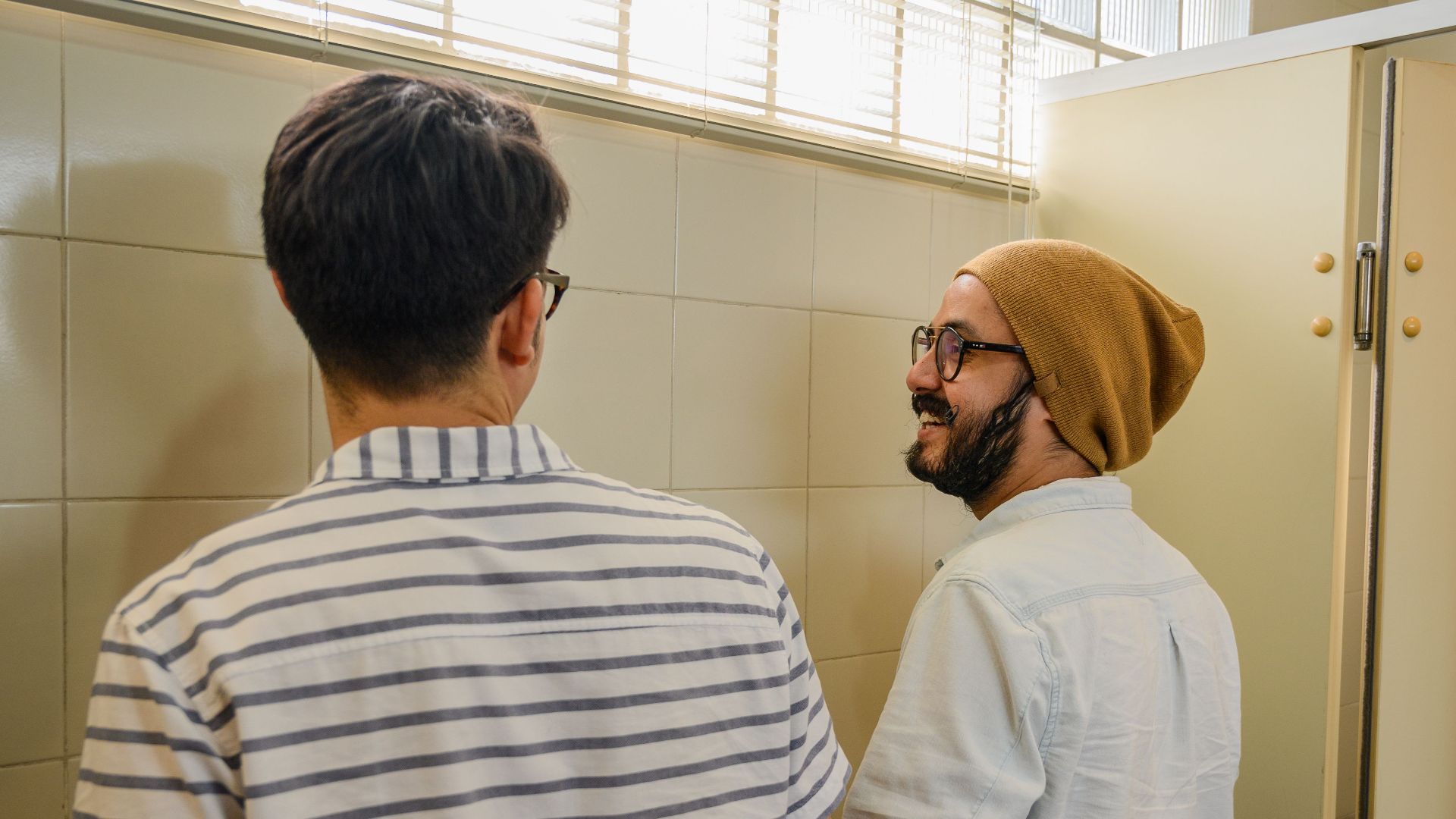 Two men wearing casual attire having a conversation in a restroom at urinals.