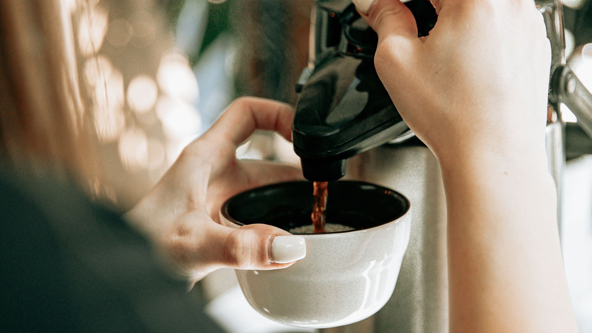 Close-up of a woman pouring coffee outdoors with a bright, bokeh background.