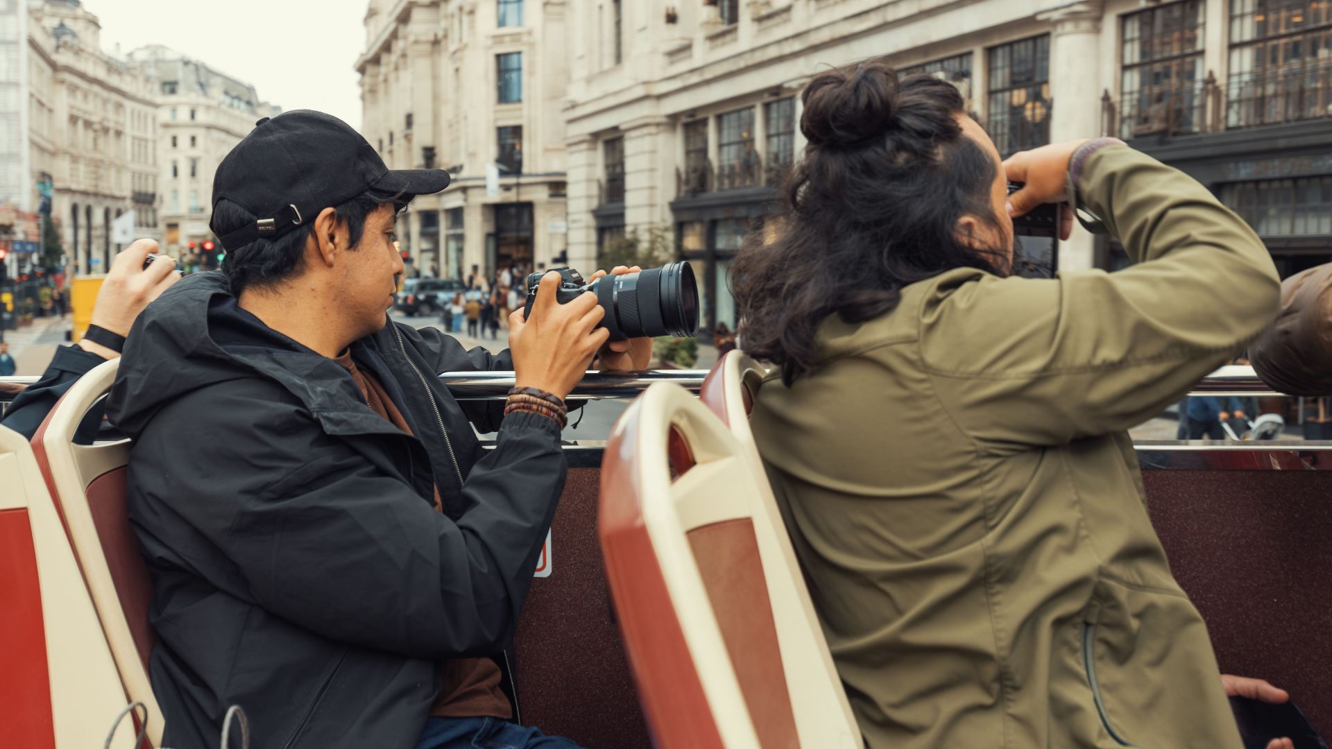 Travelers on an open top bus take photos of bustling city streets and architecture.