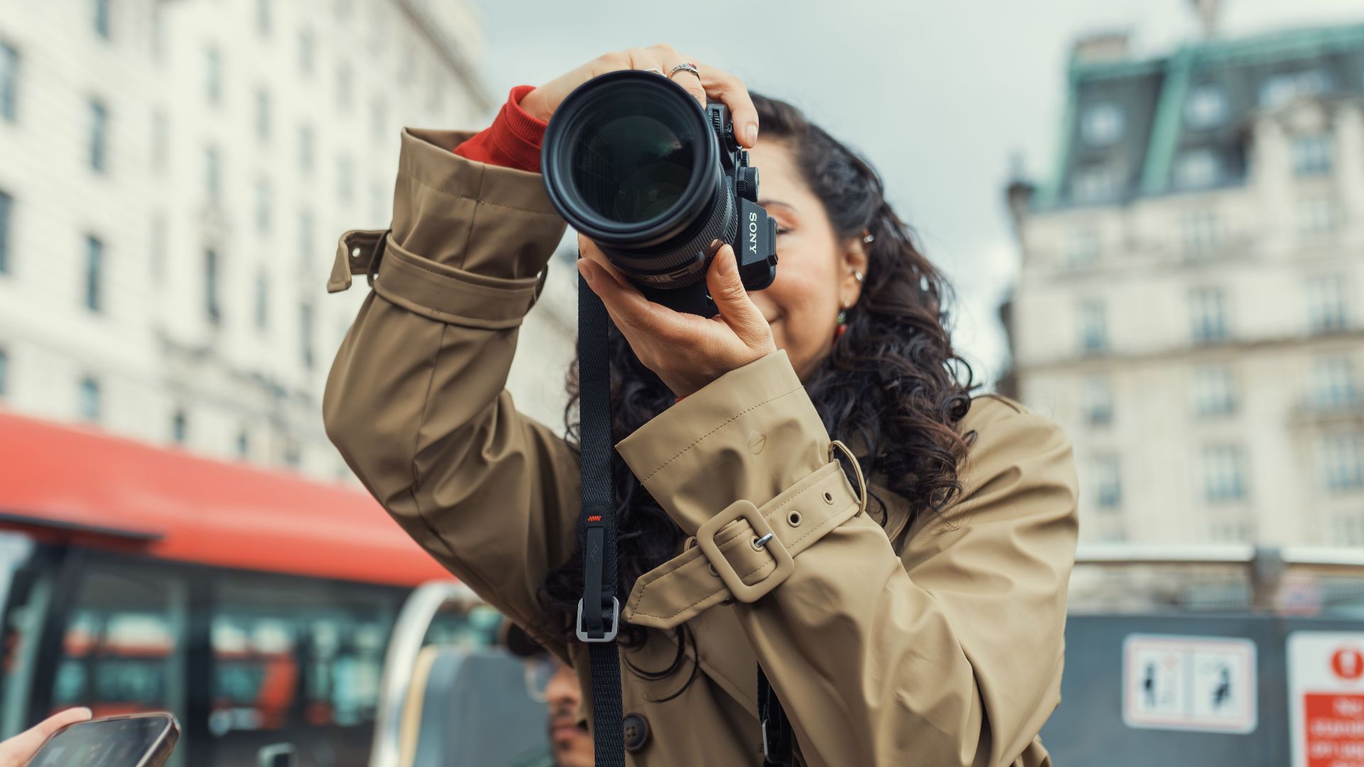 A woman with a camera takes photos from an open-top city tour bus in an urban setting.