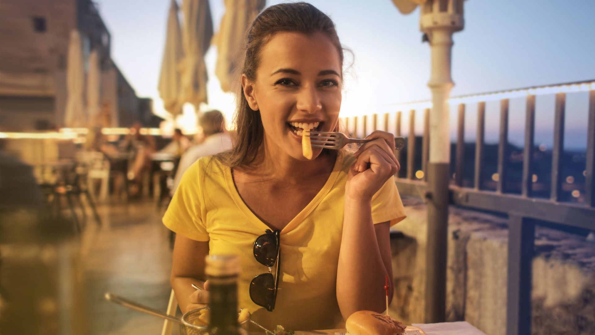 Woman in yellow top smiling while dining on a burger at a sunset outdoor cafe.