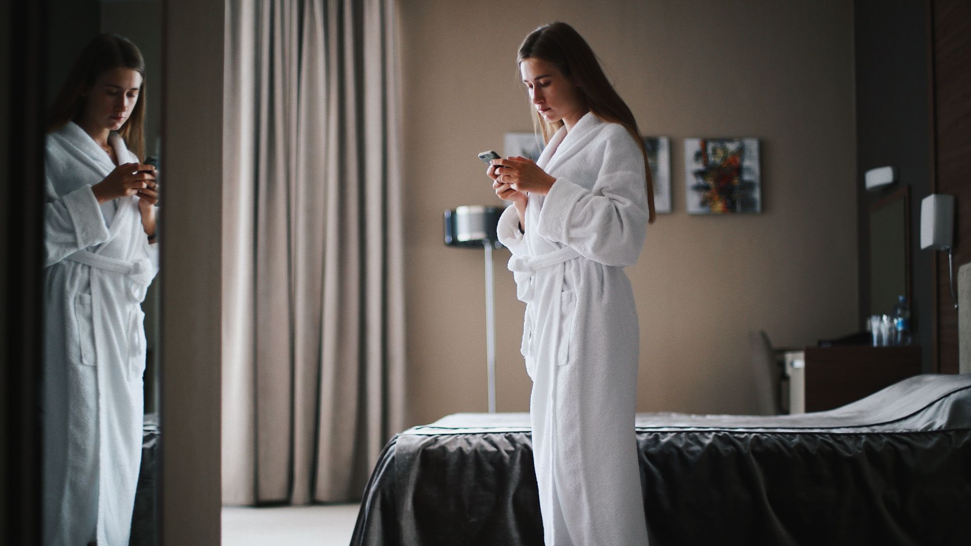 Woman in bathrobe looking at smartphone in a modern hotel room, near a mirror.