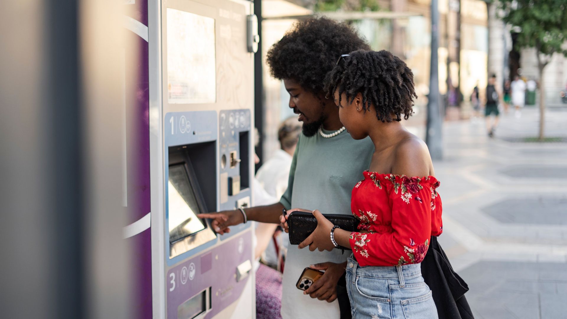 A young couple buying tickets using a touchscreen machine at an urban bus stop on a sunny day.
