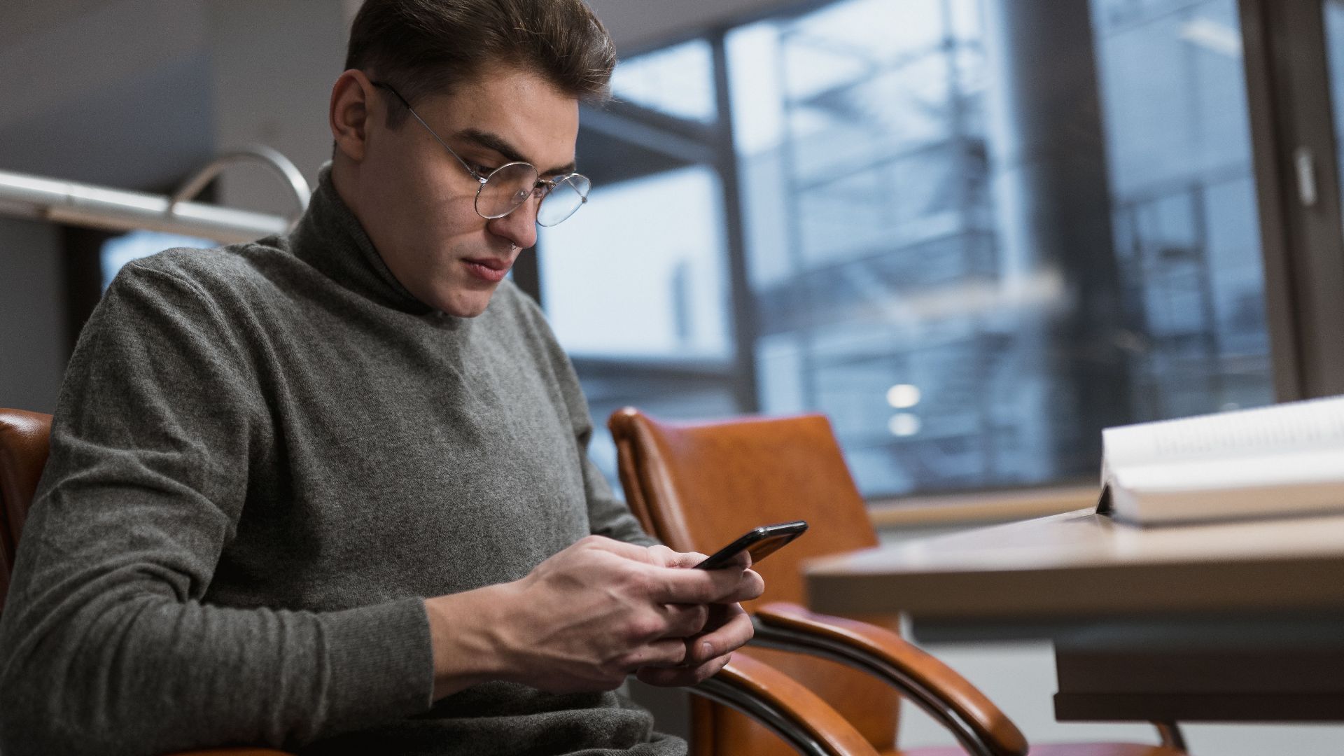 A young man in a grey sweater using a smartphone at an office desk.