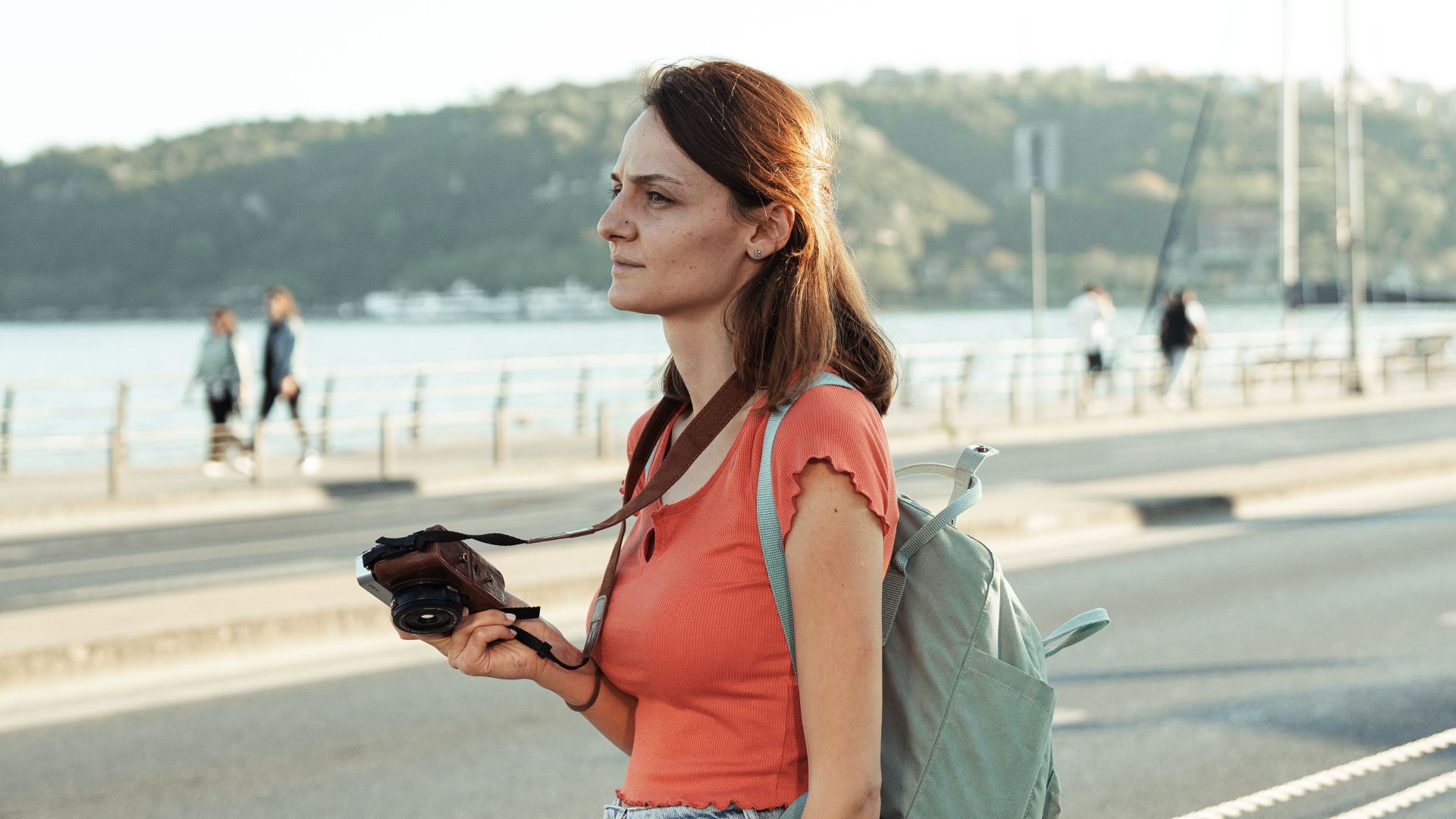 Woman standing by the waterfront with a camera and backpack on a sunny day.