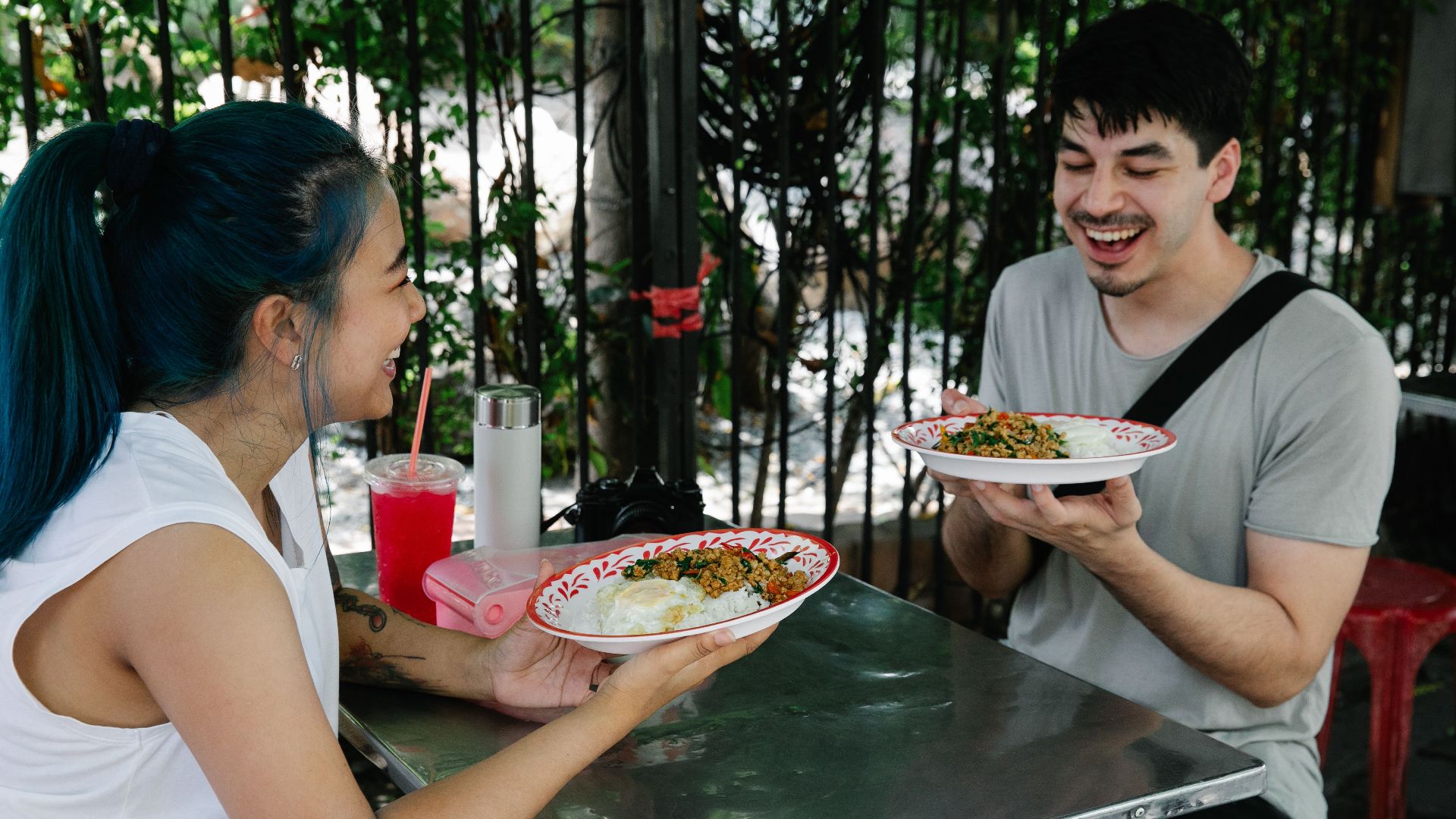 Happy multiracial couple sitting at table with plates of delicious food while having lunch together in outdoor cafeteria on summer day