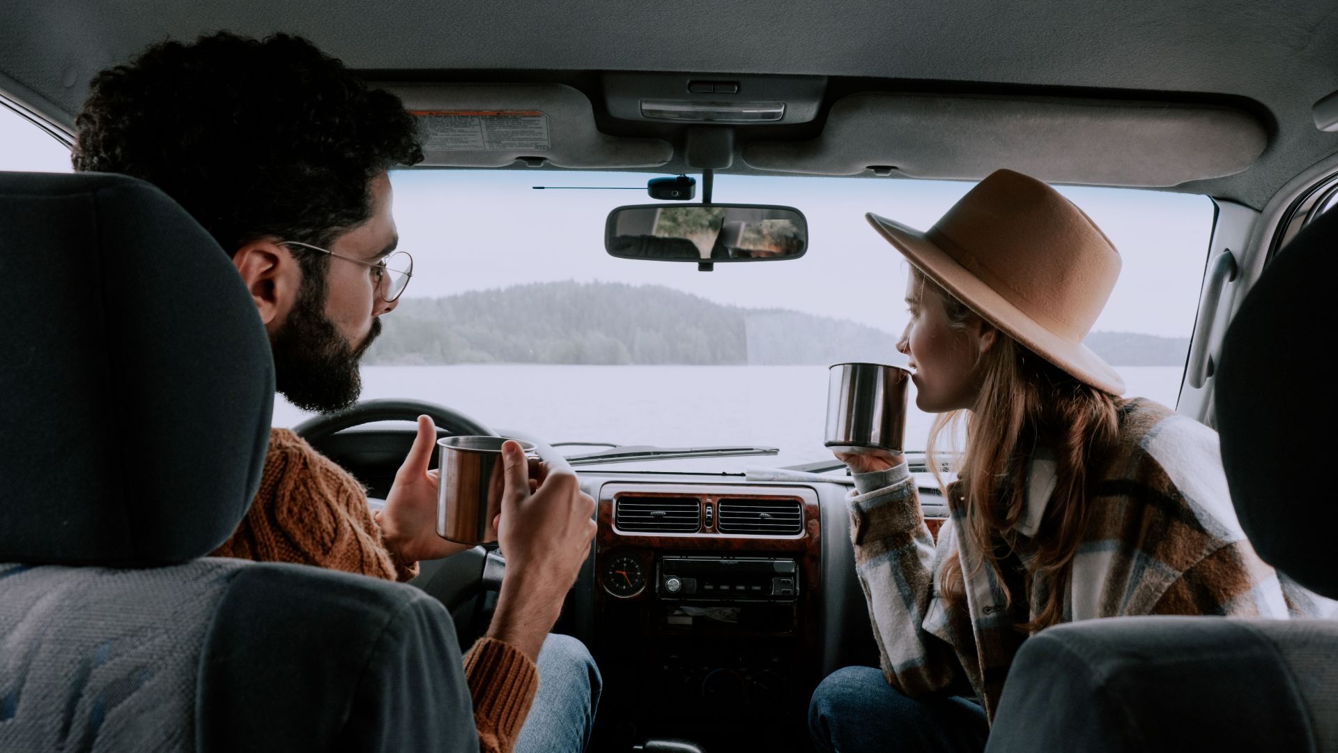 Couple enjoying hot drinks inside car with scenic lake view. Perfect for travel and lifestyle themes.