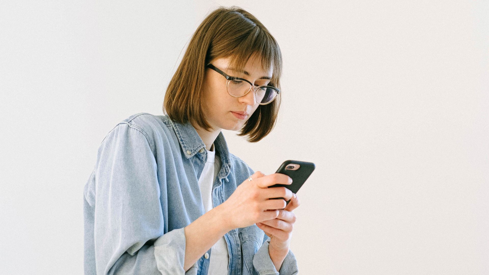 A woman in casual attire is focused on her smartphone in a minimalist home office setting.