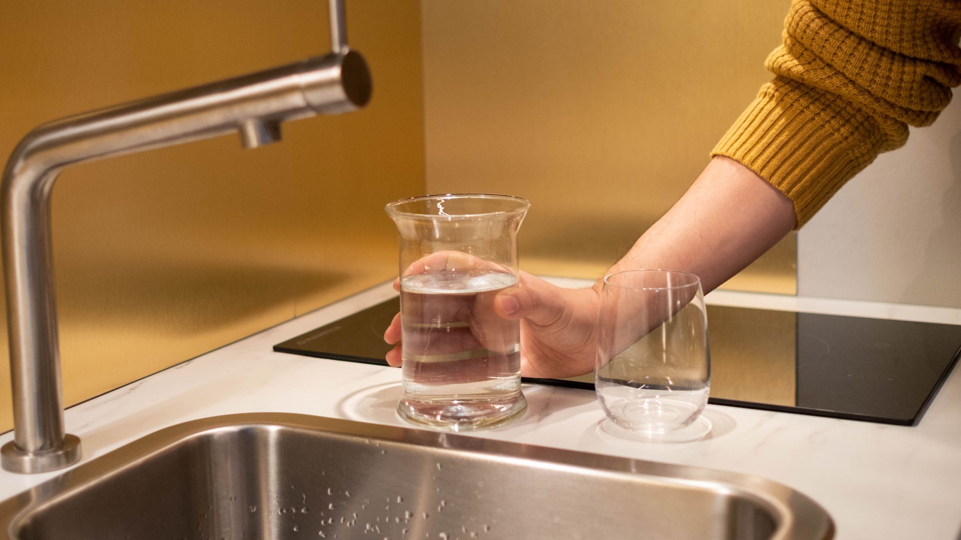 A person in a yellow sweater fills a glass of water at a modern kitchen sink.
