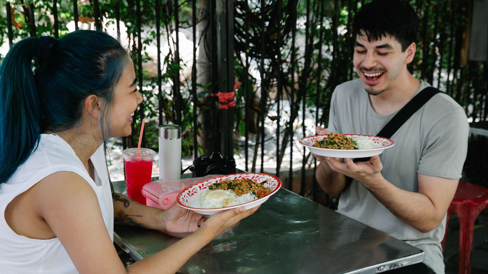 Happy multiracial couple sitting at table with plates of delicious food while having lunch together in outdoor cafeteria on summer day
