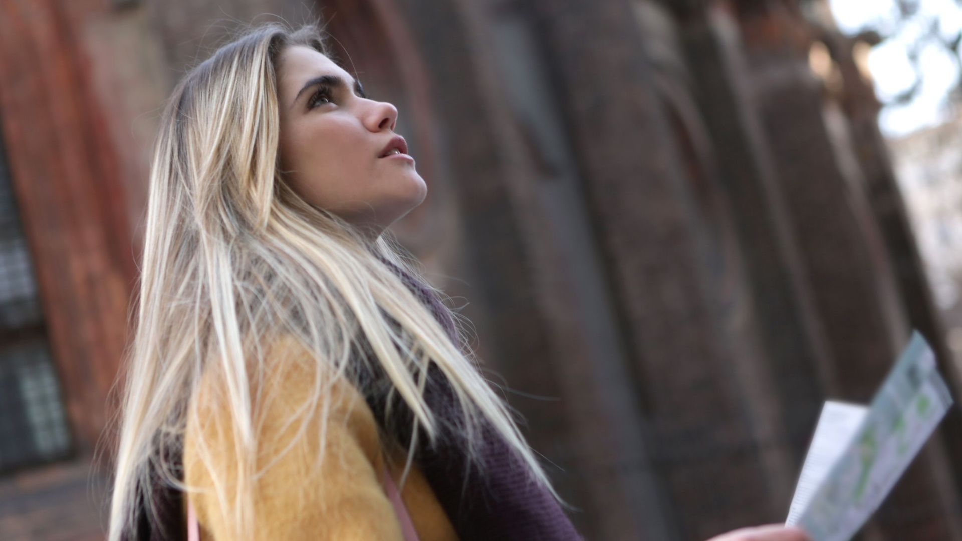 A young woman with long hair looks up while holding a map in an urban setting.