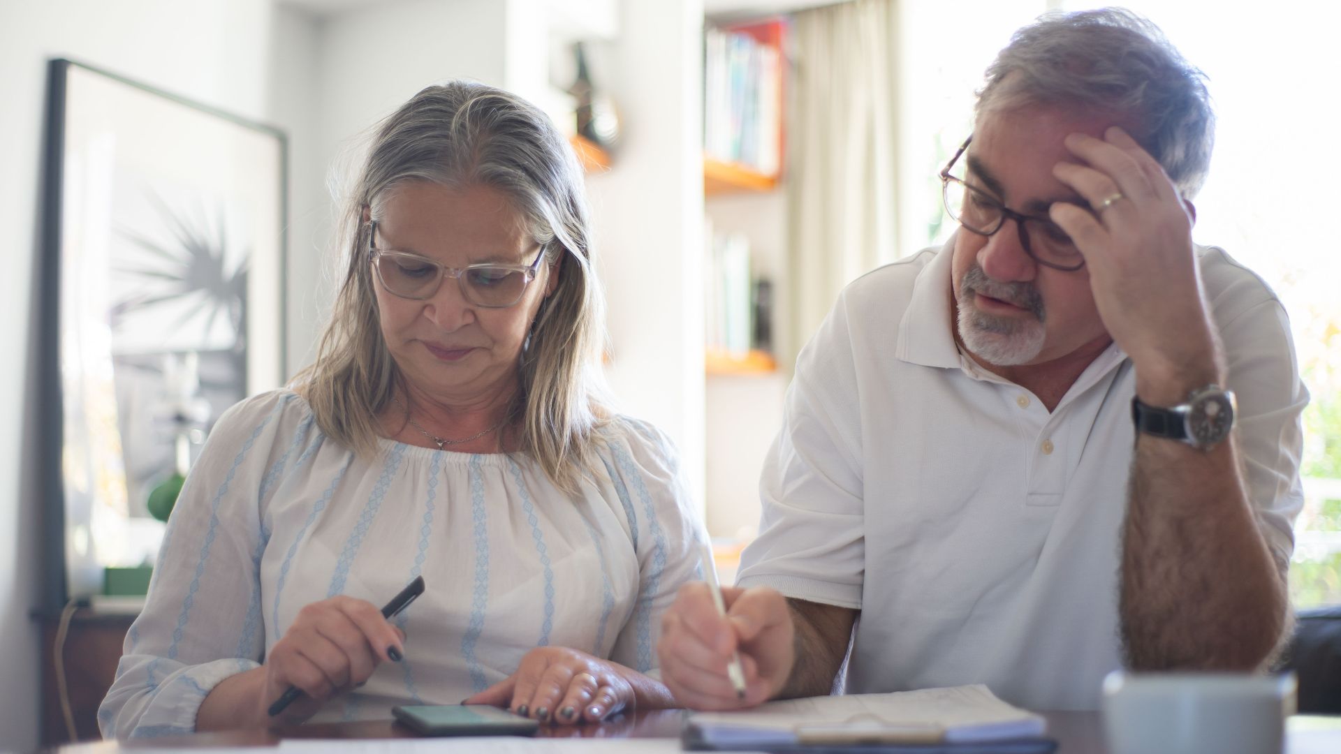 Senior couple calculating expenses at home office desk with documents and notes.
