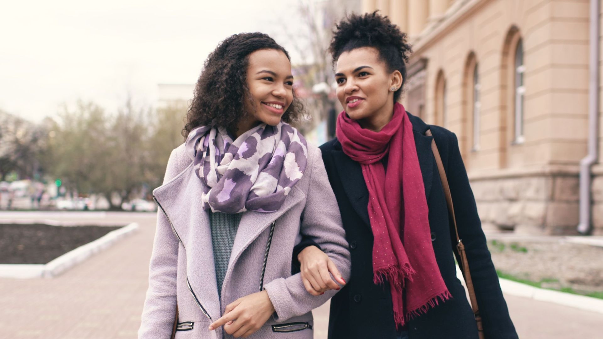 Two women joyfully carrying shopping bags while walking arm in arm in an urban setting.