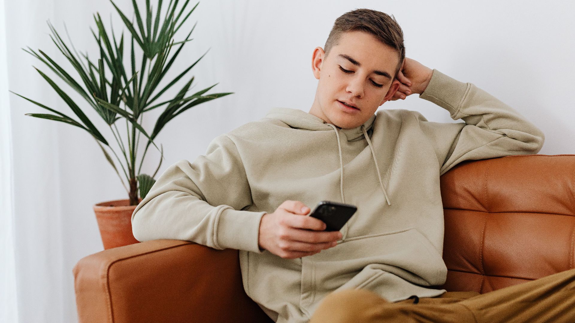 Teenage boy sitting indoors texting on a smartphone, wearing a hoodie.