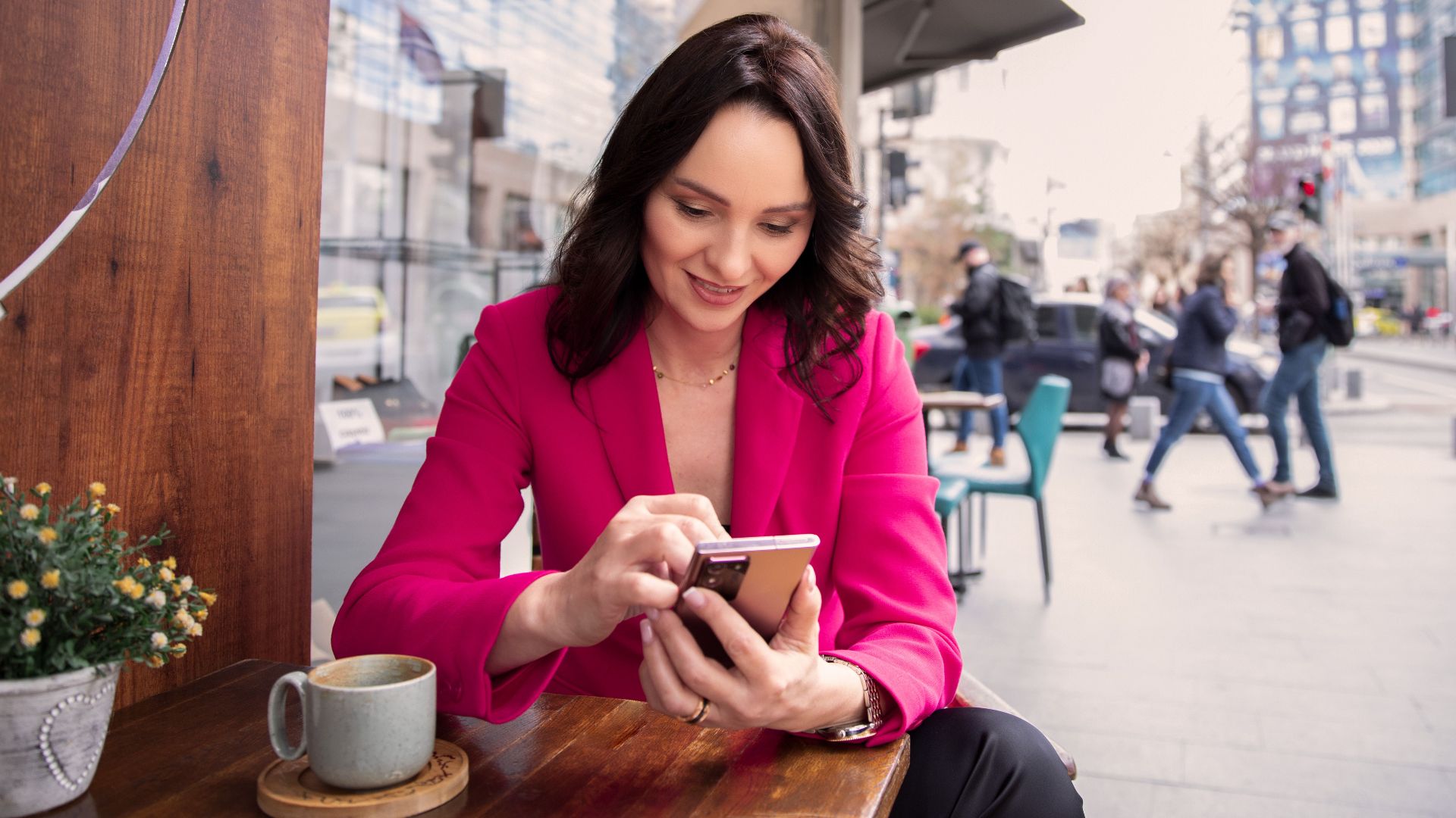 A businesswoman in a pink jacket uses her smartphone at a café table in an urban setting.