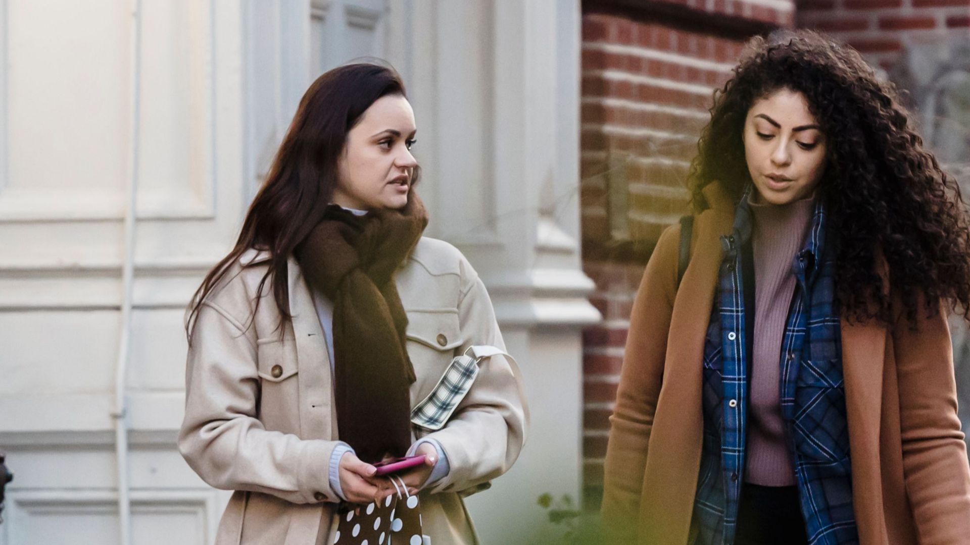 Full body of pensive young diverse women with long dark hair in casual outfits strolling on sidewalk near buildings and chatting after shopping in city