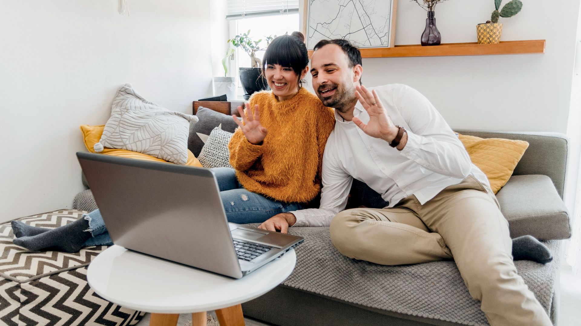 Happy couple sitting on couch, using laptop for a video call in a stylish living room.