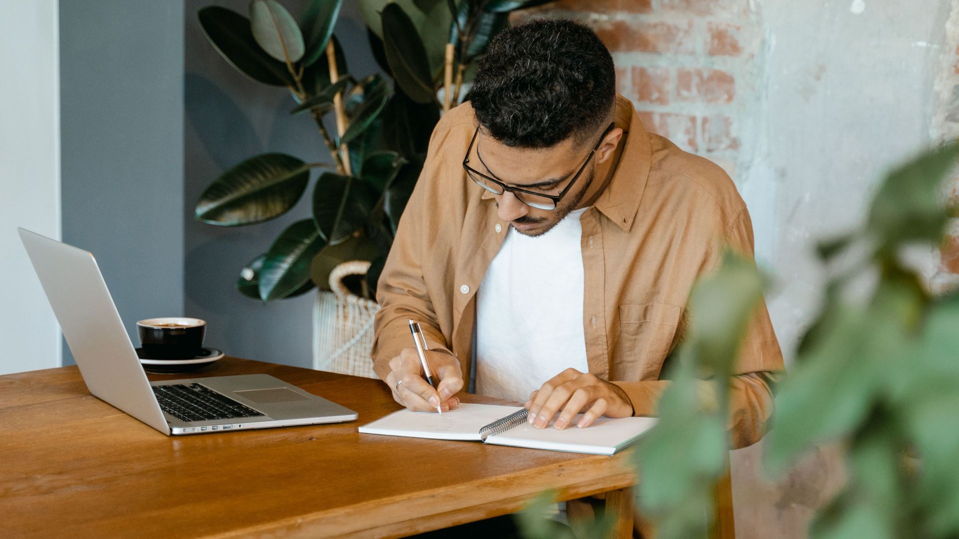 A young man concentrating at a home workspace, taking notes beside a laptop.