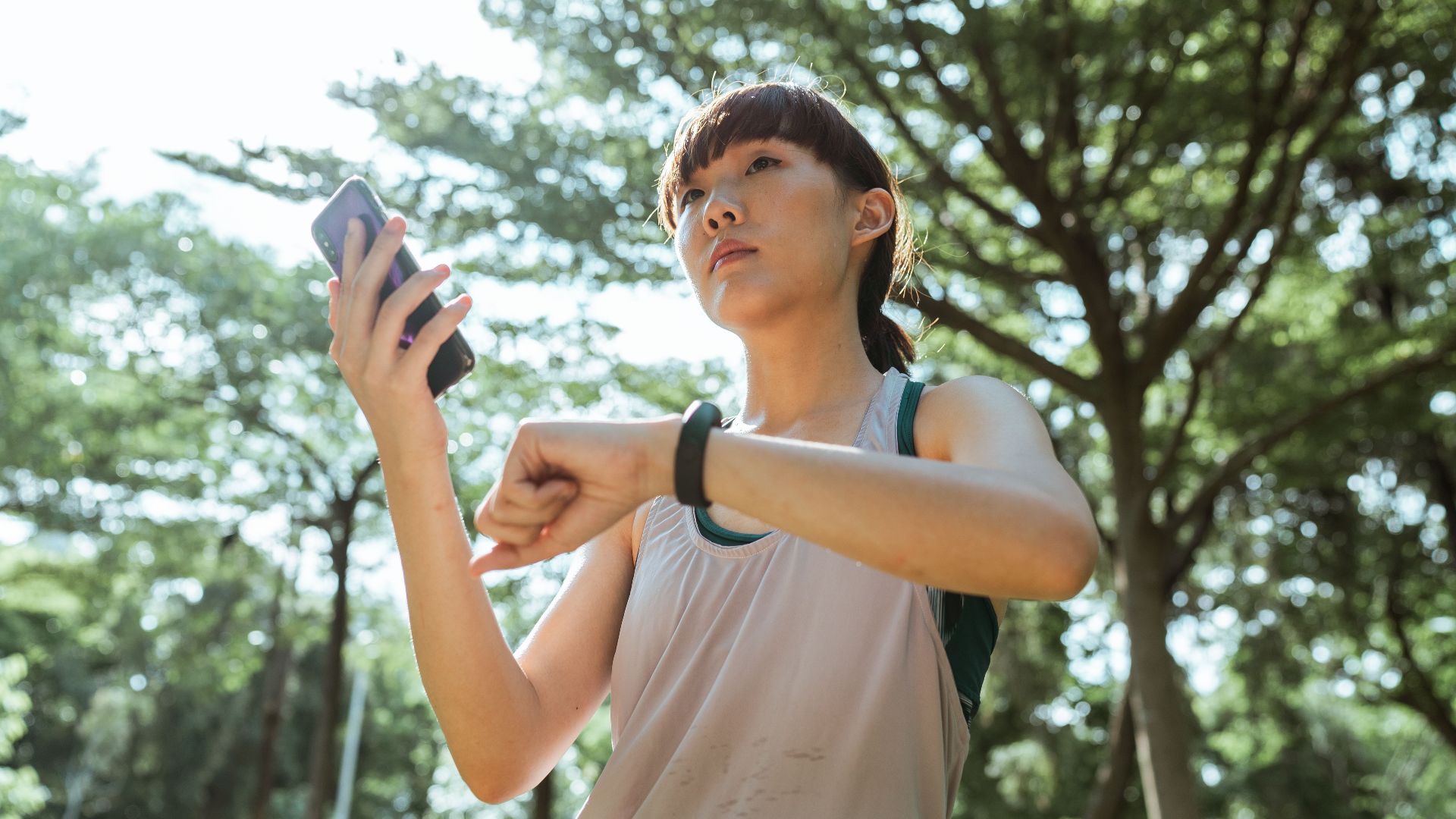 From below ethnic Asian female wearing sport clothes standing in park and connecting smartphone and smart watch during break in sunny day