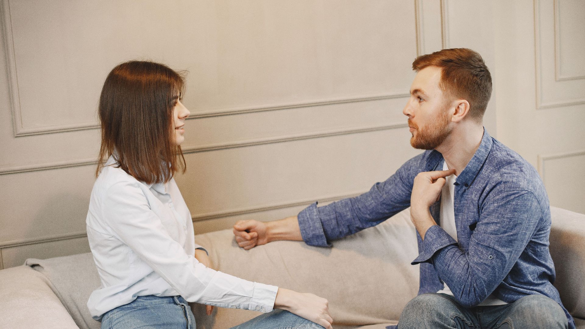 A young couple engaged in a conversation while sitting on a beige couch indoors.