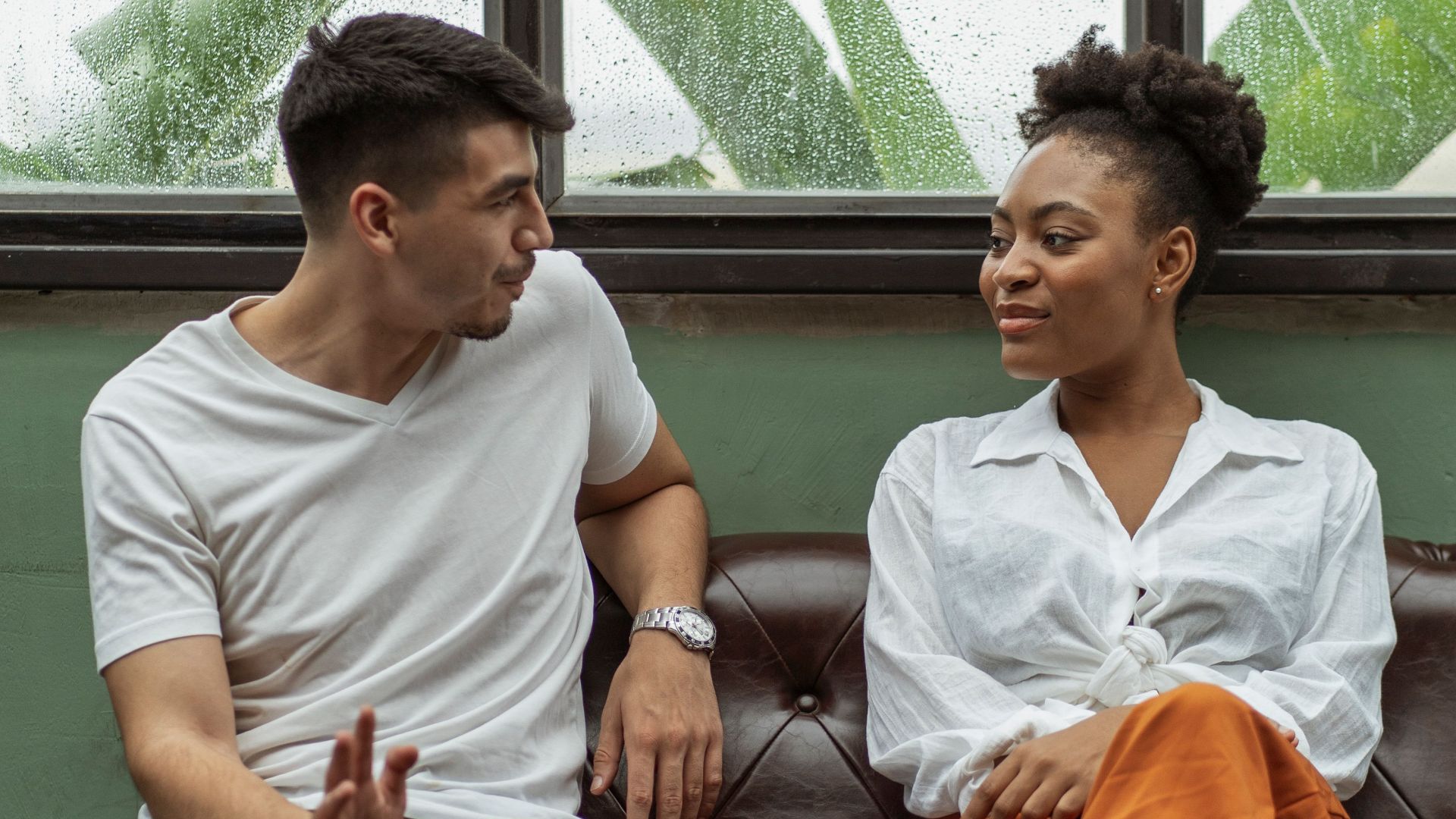 Two adults having a casual conversation on a leather couch indoors.