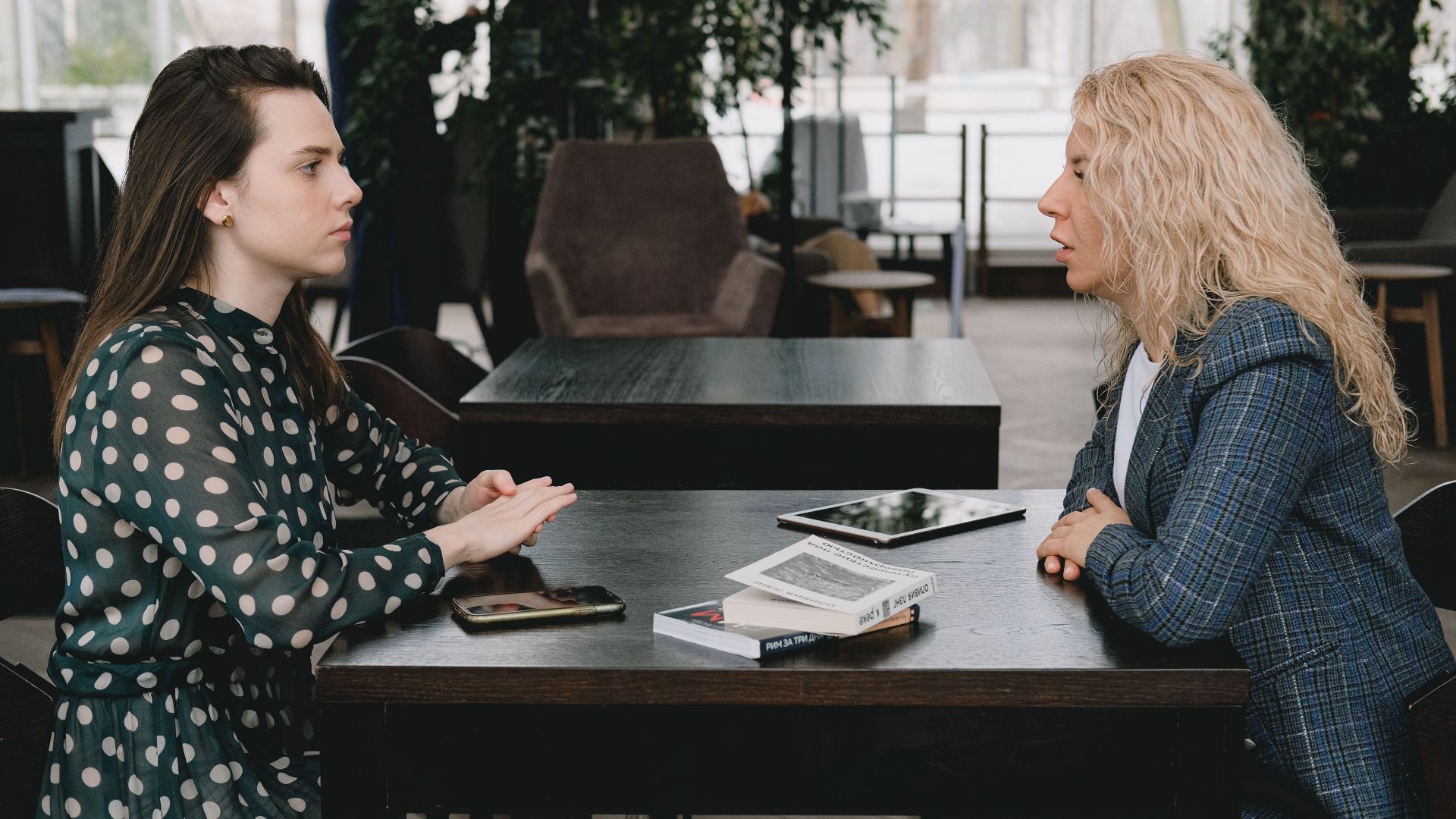 Side view of young concentrated female coworkers sitting at table with gadgets and books in modern cafe and discussing business strategy during break