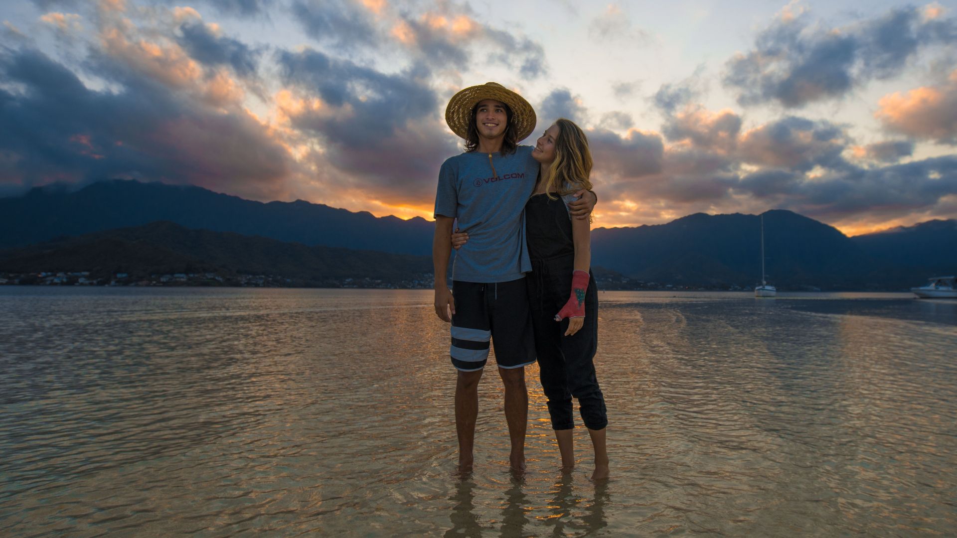 A couple shares a romantic moment on Kaneohe Beach during a picturesque sunset, surrounded by ocean and mountains.