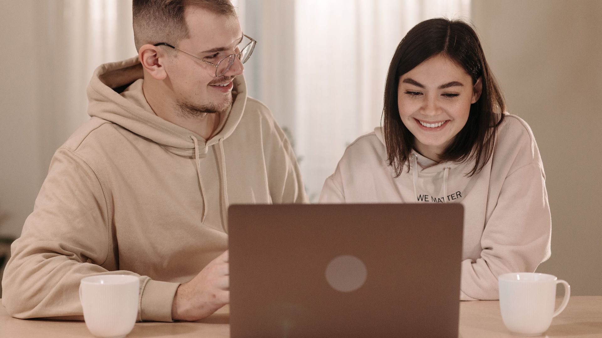 A happy couple in hoodies working together on a laptop at home, enjoying hot drinks.