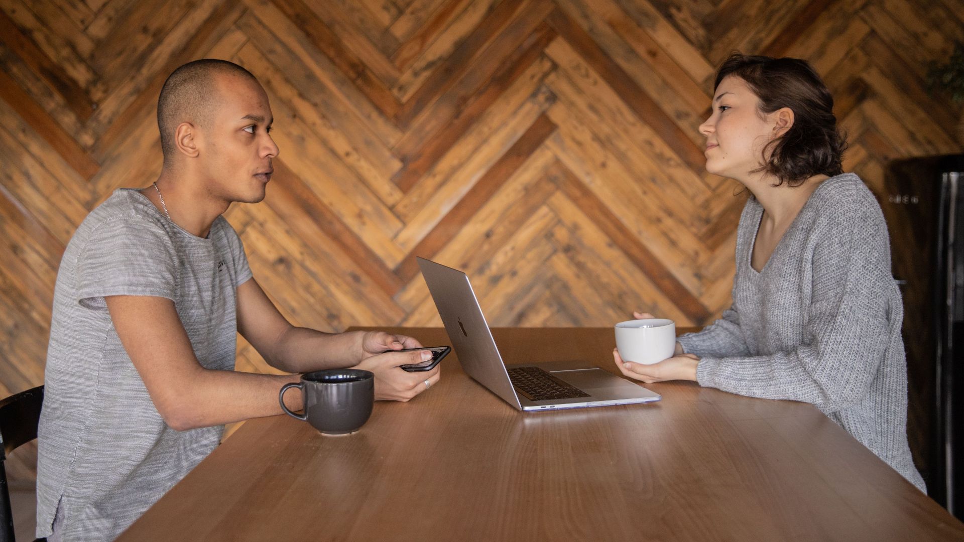 Young couple of colleagues sitting at wooden table with laptop and drinking coffee while talking about business strategy during business appointment at home looking at each other