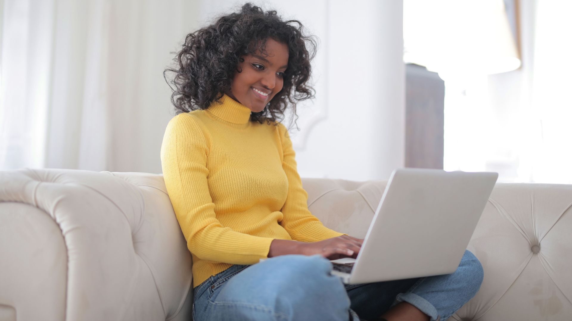 Positive young ethnic lady in casual yellow sweater and jeans sitting on comfortable couch and working on laptop while spending time at home