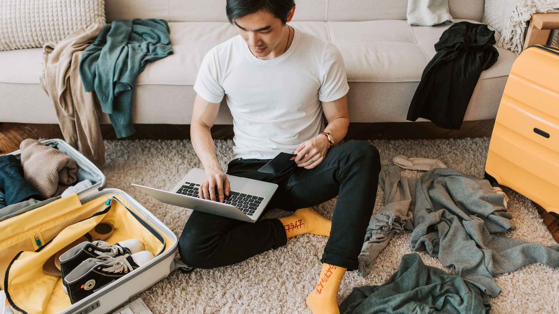 Man multitasking by packing luggage while working on a laptop in living room.