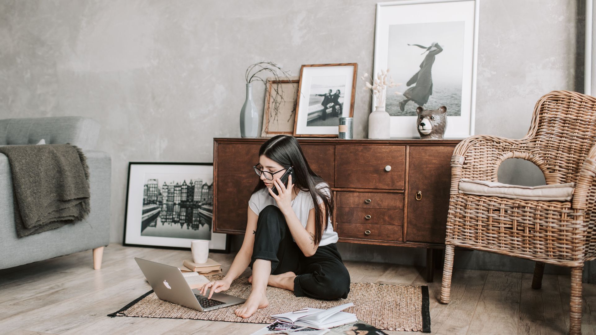 Concentrated young barefoot woman in casual outfit browsing computer while typing message and talking on mobile phone during remote work at home