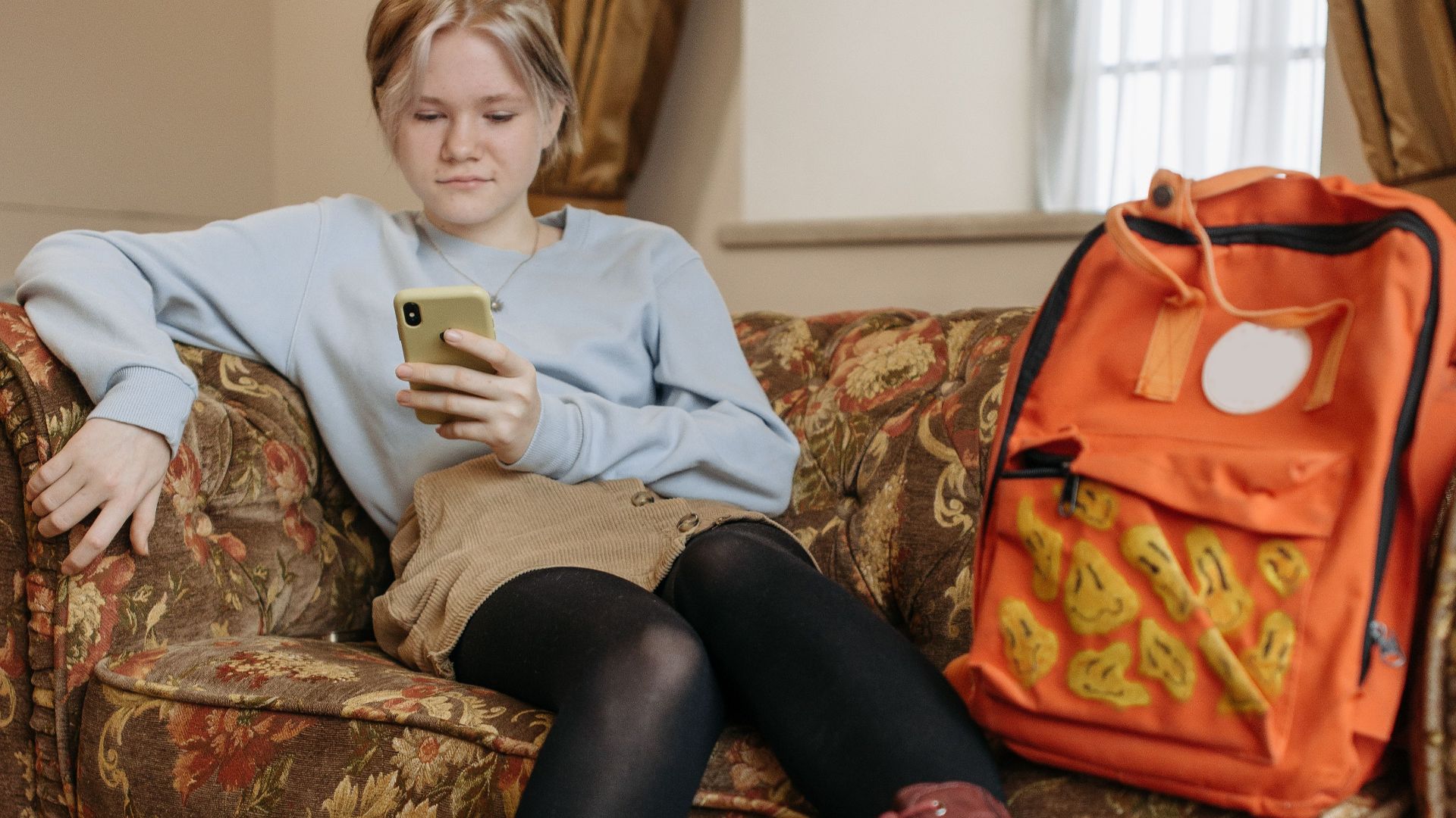 A young woman sits on a couch with a smartphone and orange backpack, looking relaxed.