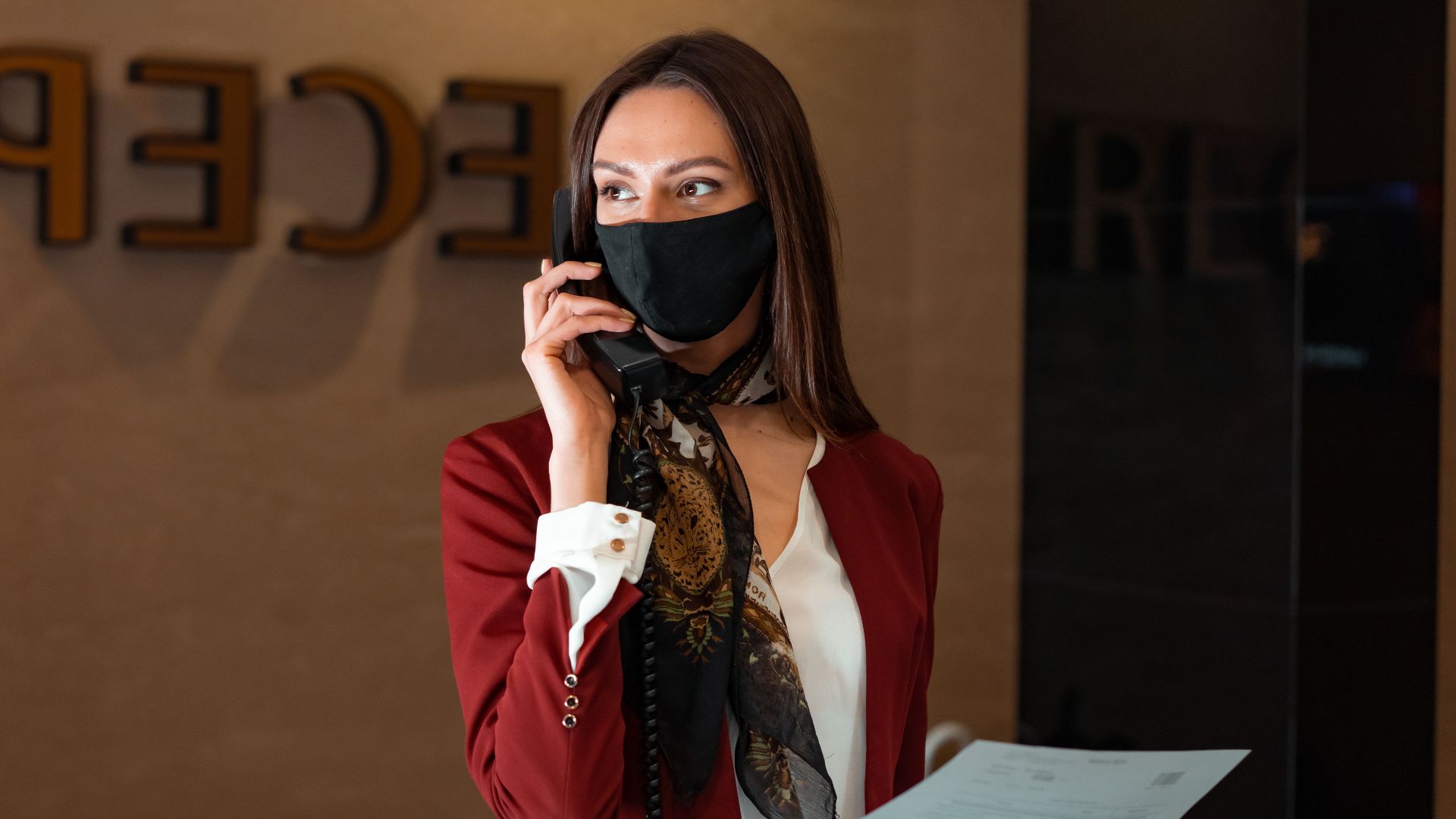 A female receptionist with a face mask answers a phone call, embodying professional service and safety.