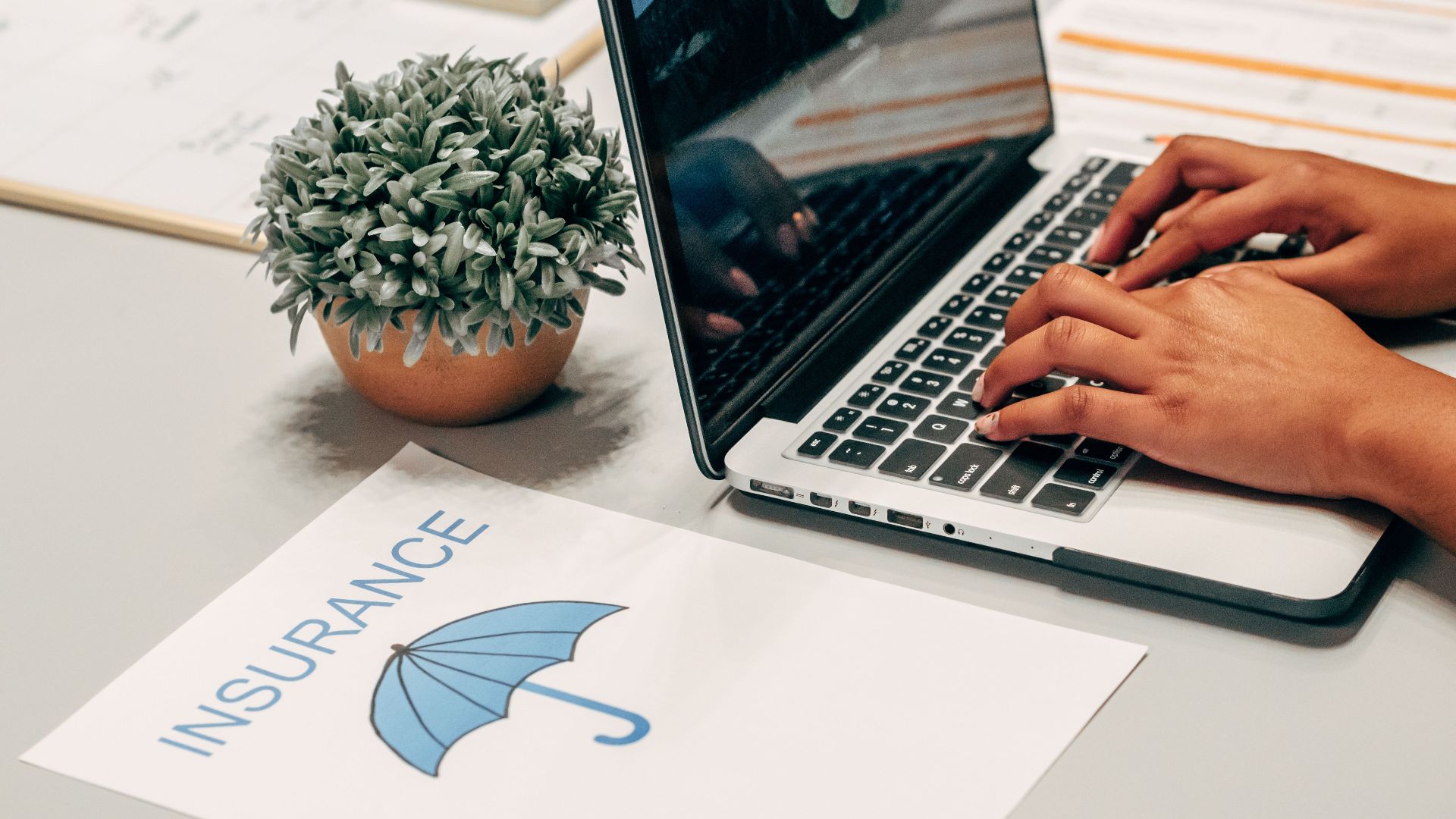 Hands typing on a laptop at a desk with an insurance paper and plant, suggesting a work environment.