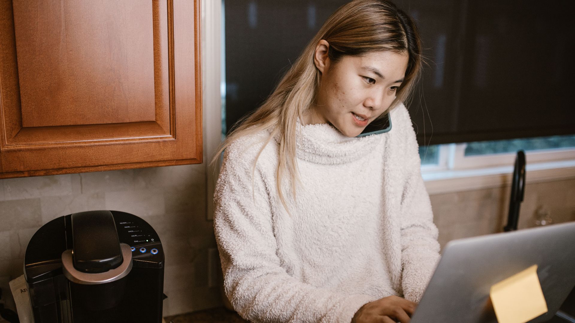 Woman multitasking on laptop and phone in a warm kitchen setup.