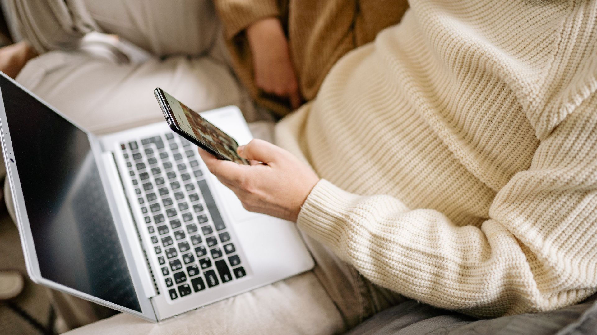Adult in a cozy sweater multitasking with a smartphone and laptop indoors.