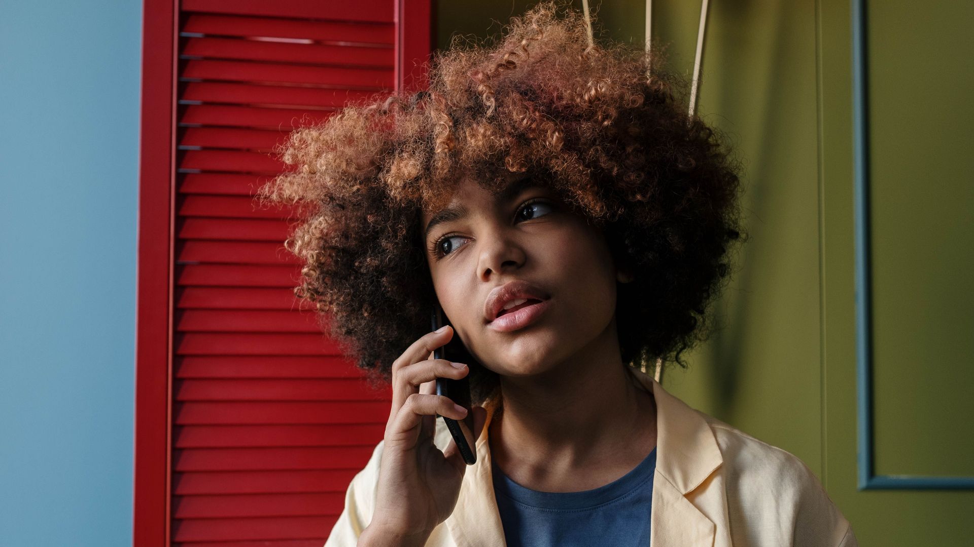 Contemplative woman with curly hair taking a phone call indoors with colorful background.
