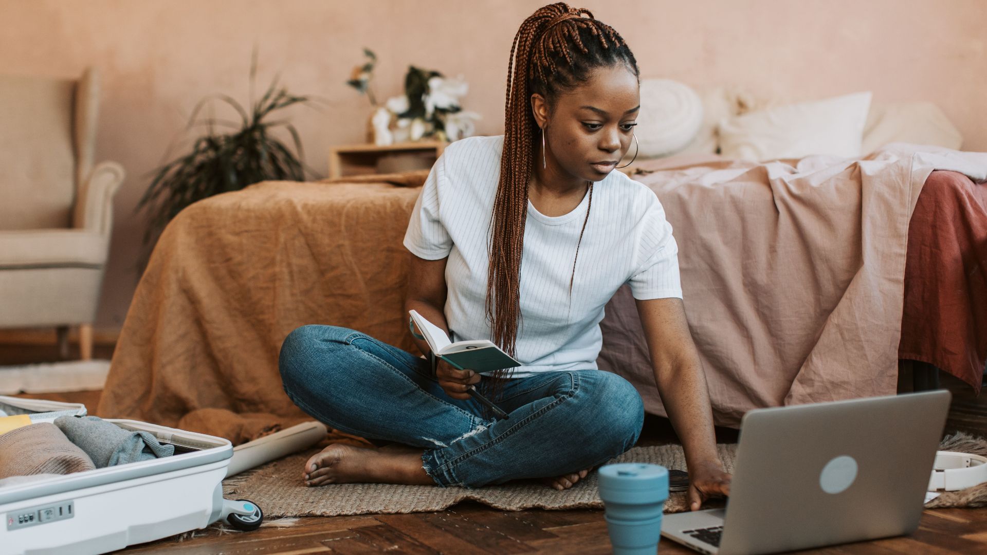 Woman sits on floor packing suitcase, using laptop, and holding notebook in a cozy bedroom environment.