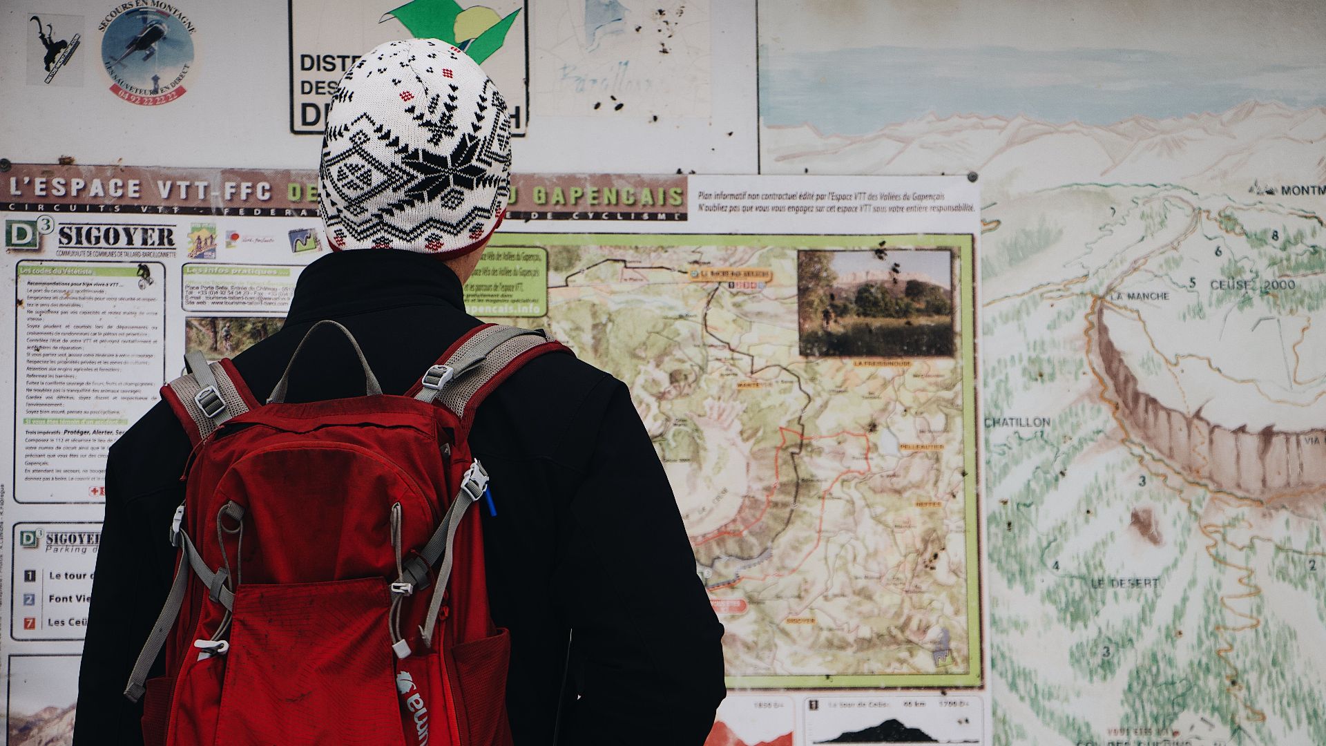 Person with a red backpack studying a detailed trail map in an outdoor mountain setting.