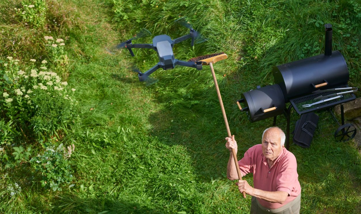 Old senior man upset by a flying drone over his garden. The concept of spying on neighbors and their privacy. Man trying to knock down a drone from the sky over his own garden with a broom. Above view