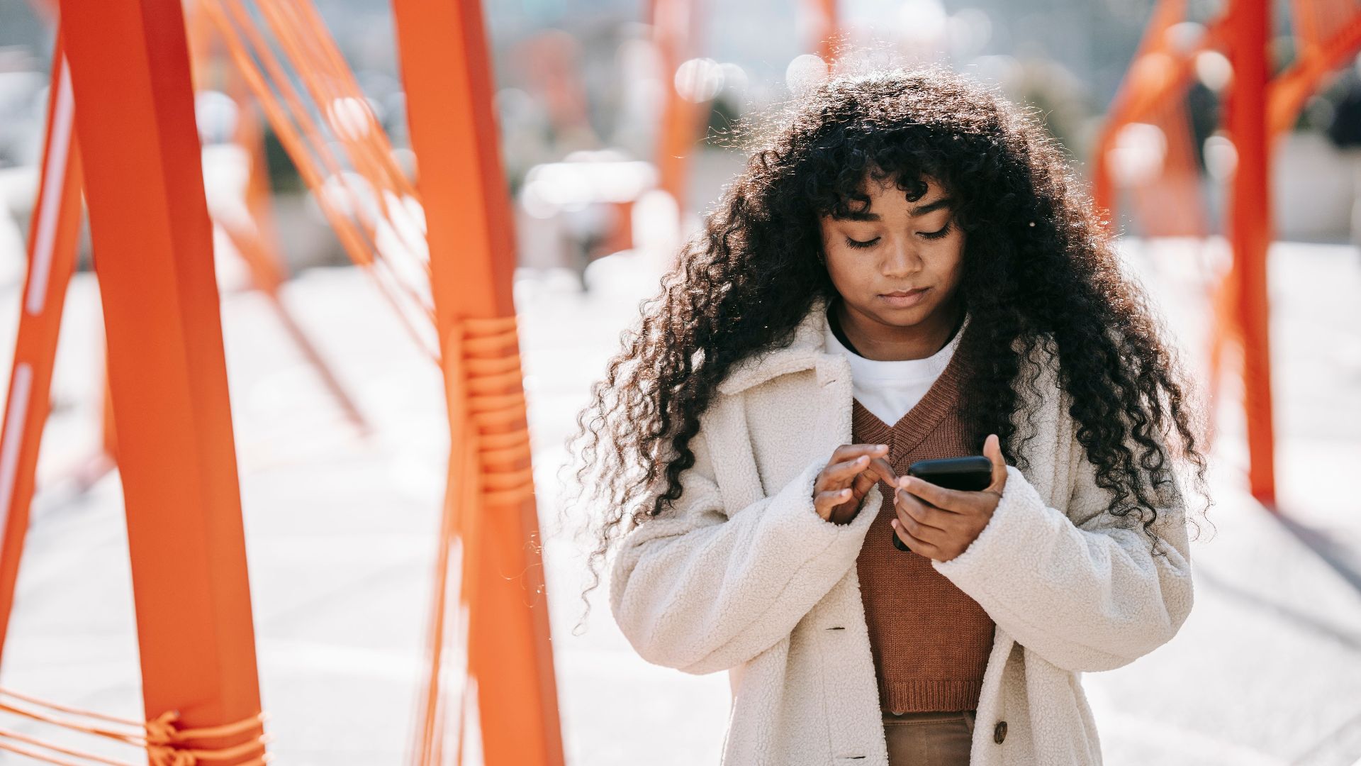 Concentrated African American female with long curly hair wearing white coat browsing mobile phone while standing on city street on sunny spring day
