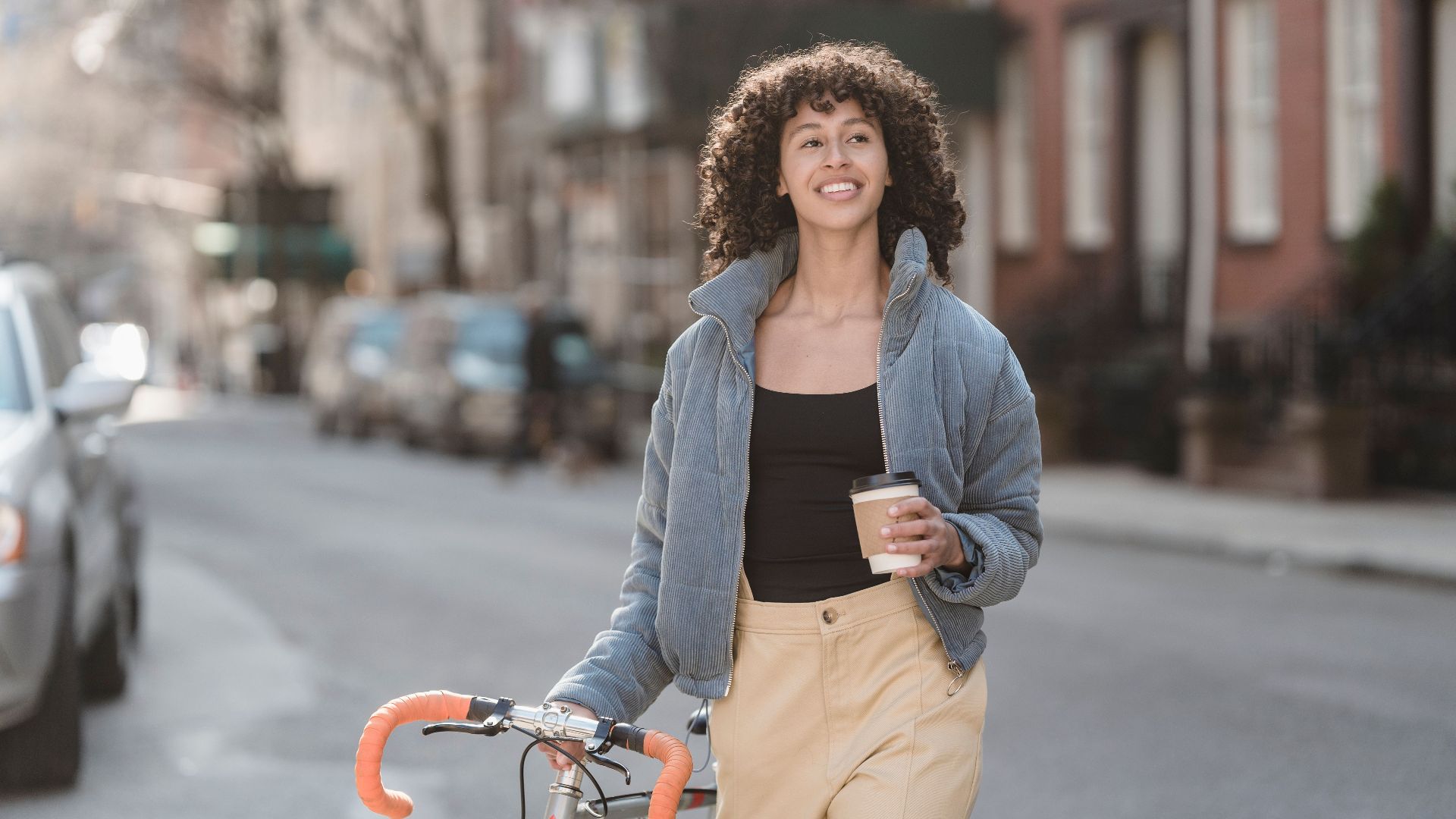 Self assured young ethnic lady with curly hair in trendy warm jacket smiling and looking away while crossing asphalt road with bicycle and cup of coffee to go in hand