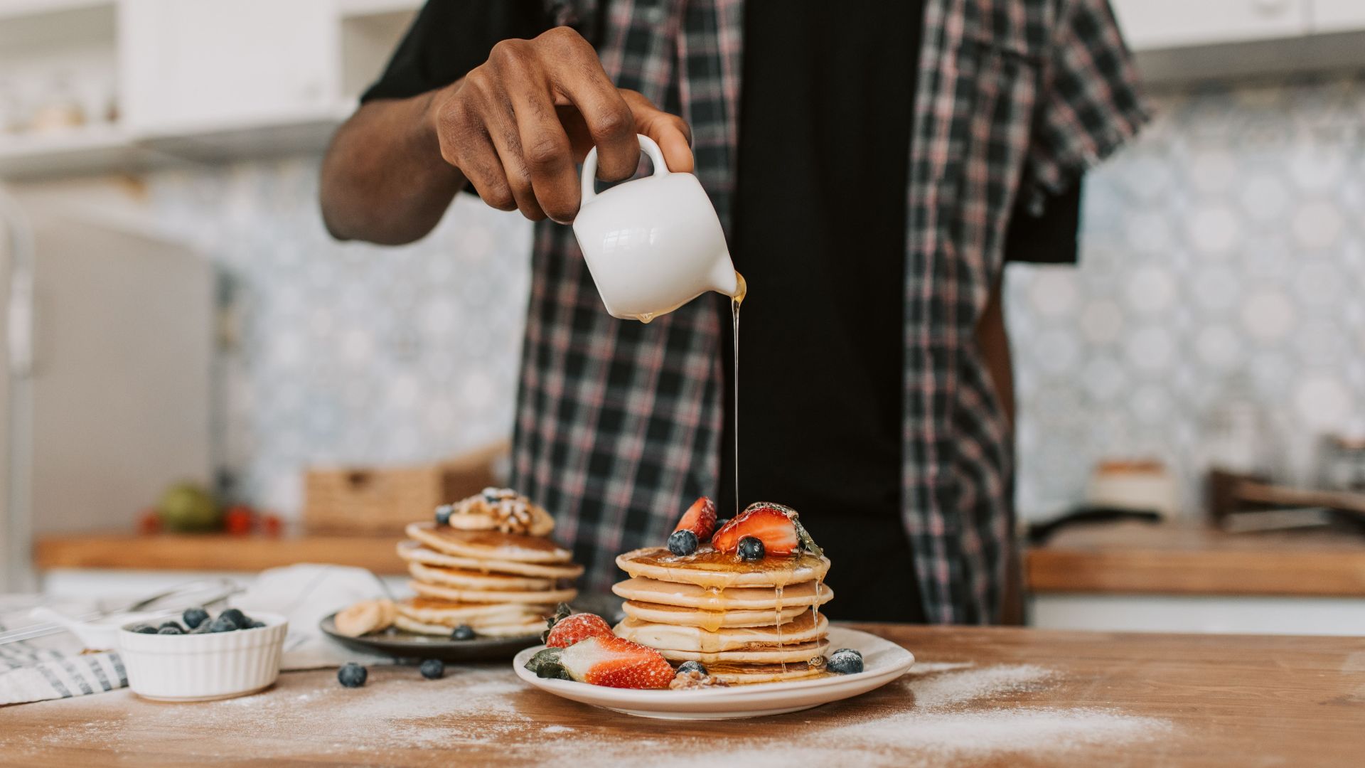 A person pouring maple syrup over a stack of pancakes topped with fresh berries.