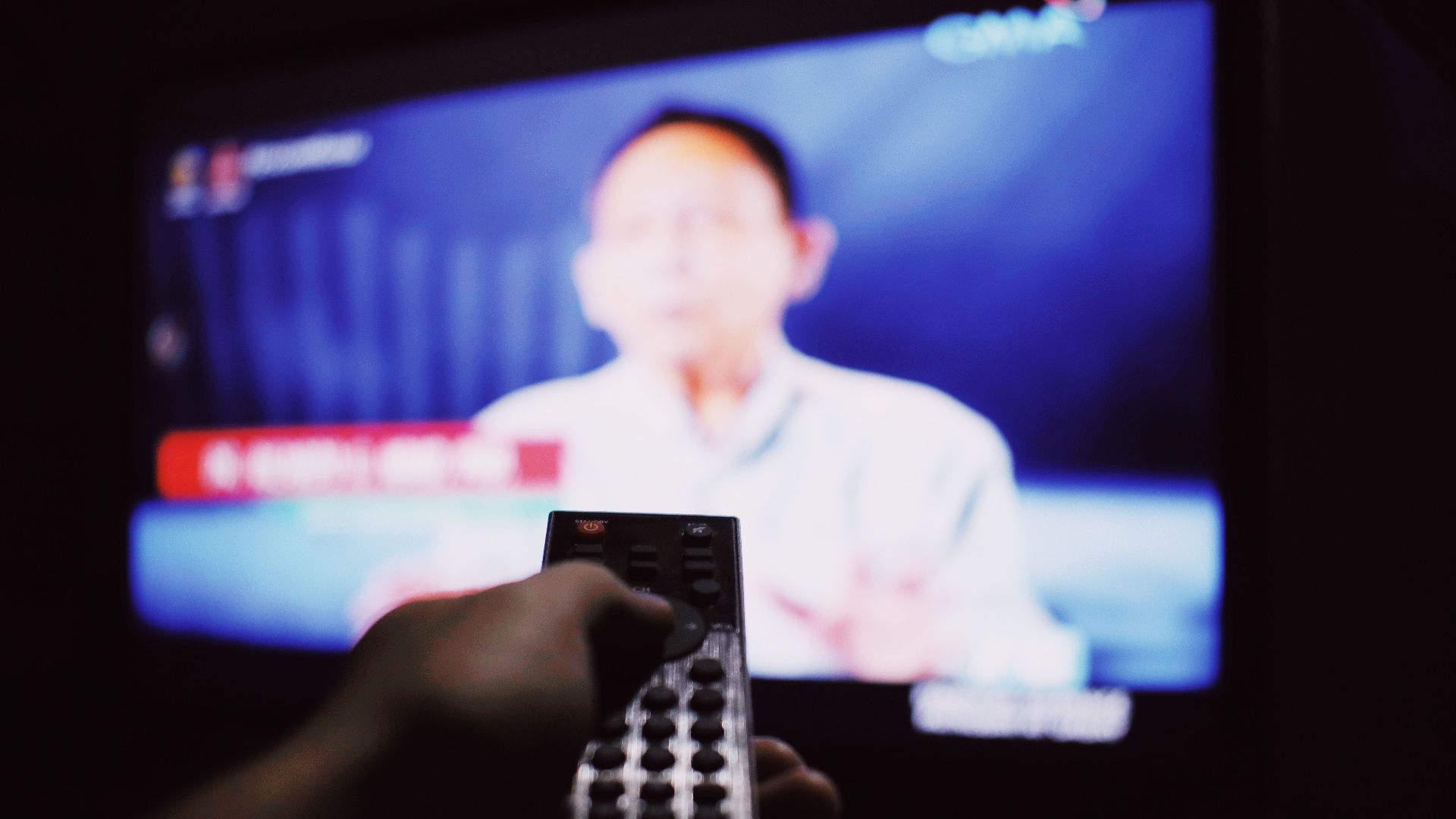 Crop anonymous person with remote controller switching channels while watching TV on blurred background in living room with dim light
